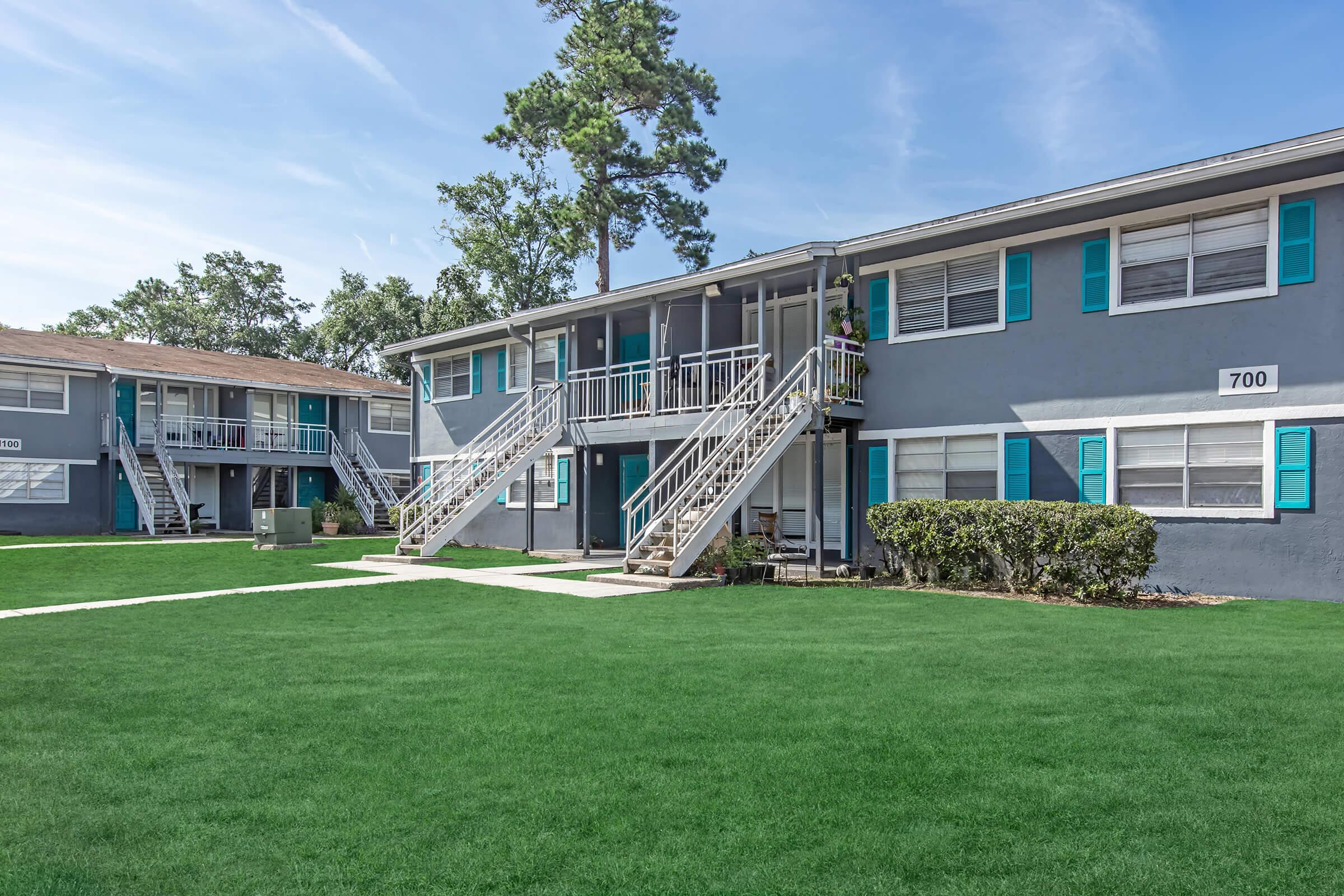 A view of a multi-unit apartment complex featuring two buildings. The buildings have gray exteriors with bright blue shutters. There are white staircases leading to the second floor, manicured lawns, and small bushes lining the pathways. The sky is clear with a few clouds, creating a pleasant atmosphere.