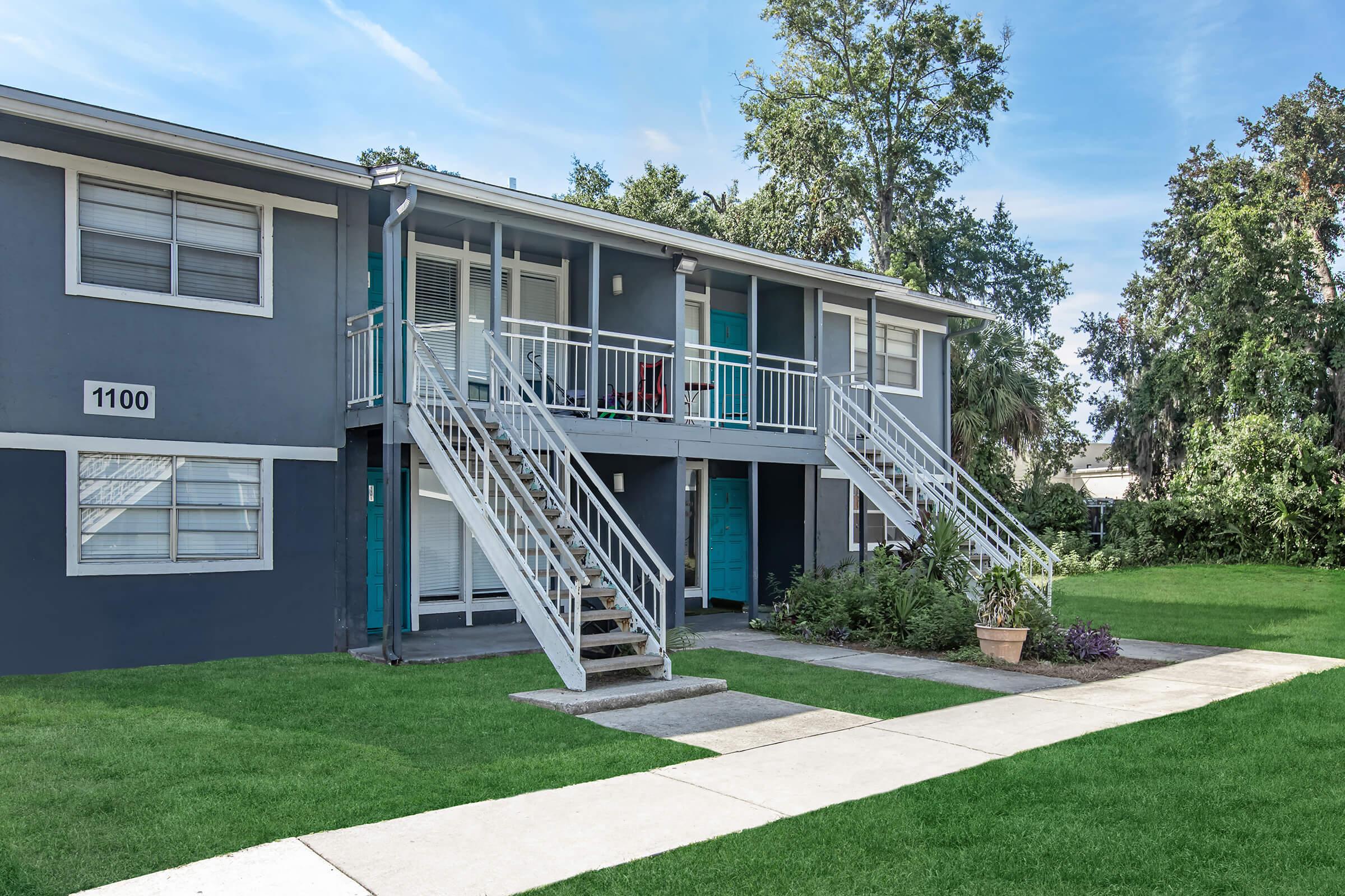 Two-story apartment building with a gray exterior featuring white railings and turquoise doors. There are stairs leading to upper units, and the landscaped yard has green grass and a potted plant. The building is set against a blue sky and surrounded by trees.
