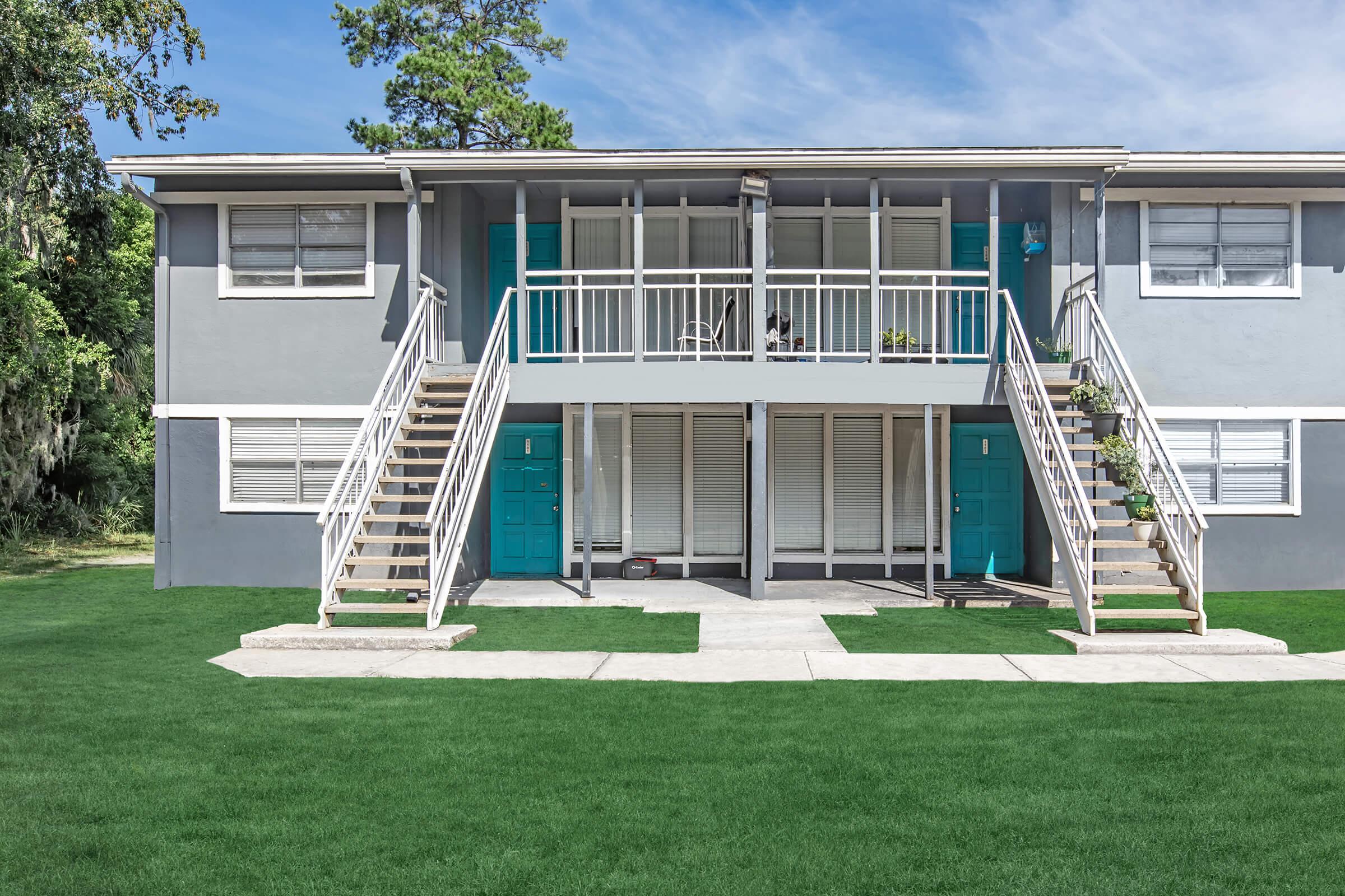 Two-level apartment building with light gray exterior and turquoise doors. The building features a set of stairs leading to each entrance, a well-maintained lawn, and several potted plants. Large windows allow ample natural light, and the sky is partly cloudy.