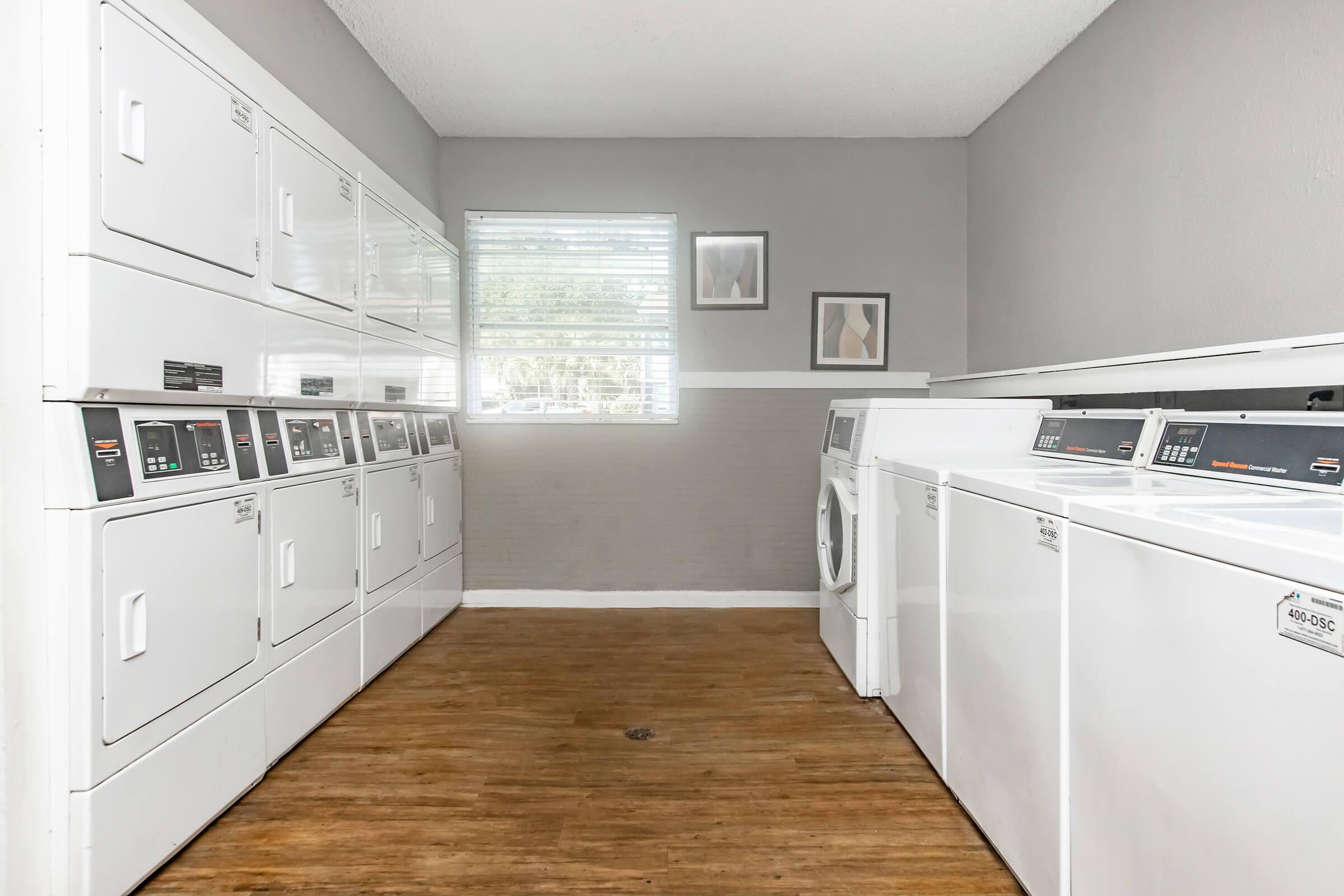 A clean and bright laundry room featuring rows of white washing machines and dryers along the right side. The left wall has stacked washers and a large window letting in natural light. The floor is wooden, and there are minimalist framed artworks on the walls, creating a modern and organized atmosphere.