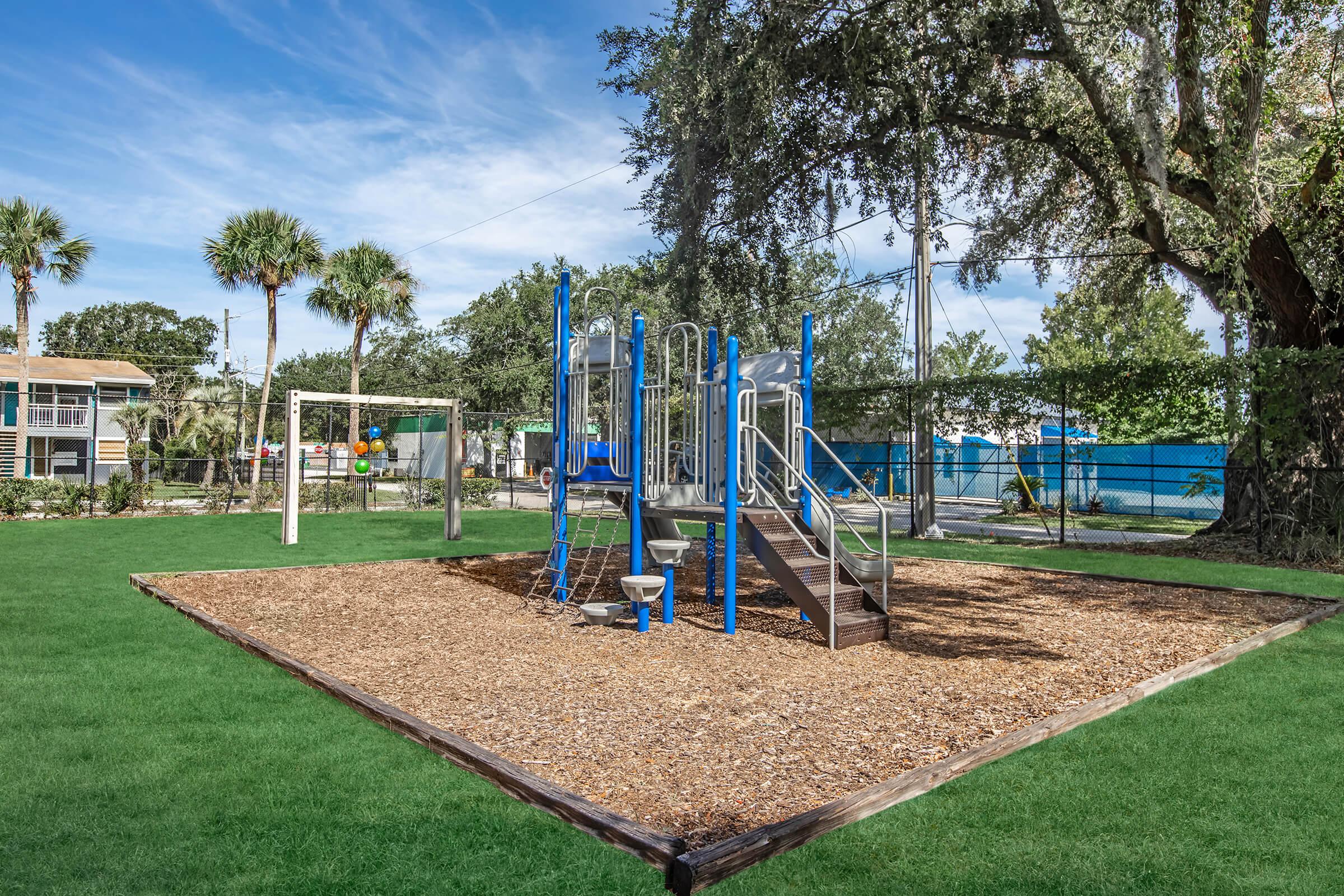 A children's playground featuring a blue climbing structure with slides, surrounded by wood chips and grassy areas. In the background, there is a swing set and palm trees, set against a clear blue sky. The playground is enclosed by a fence, providing a safe space for play.