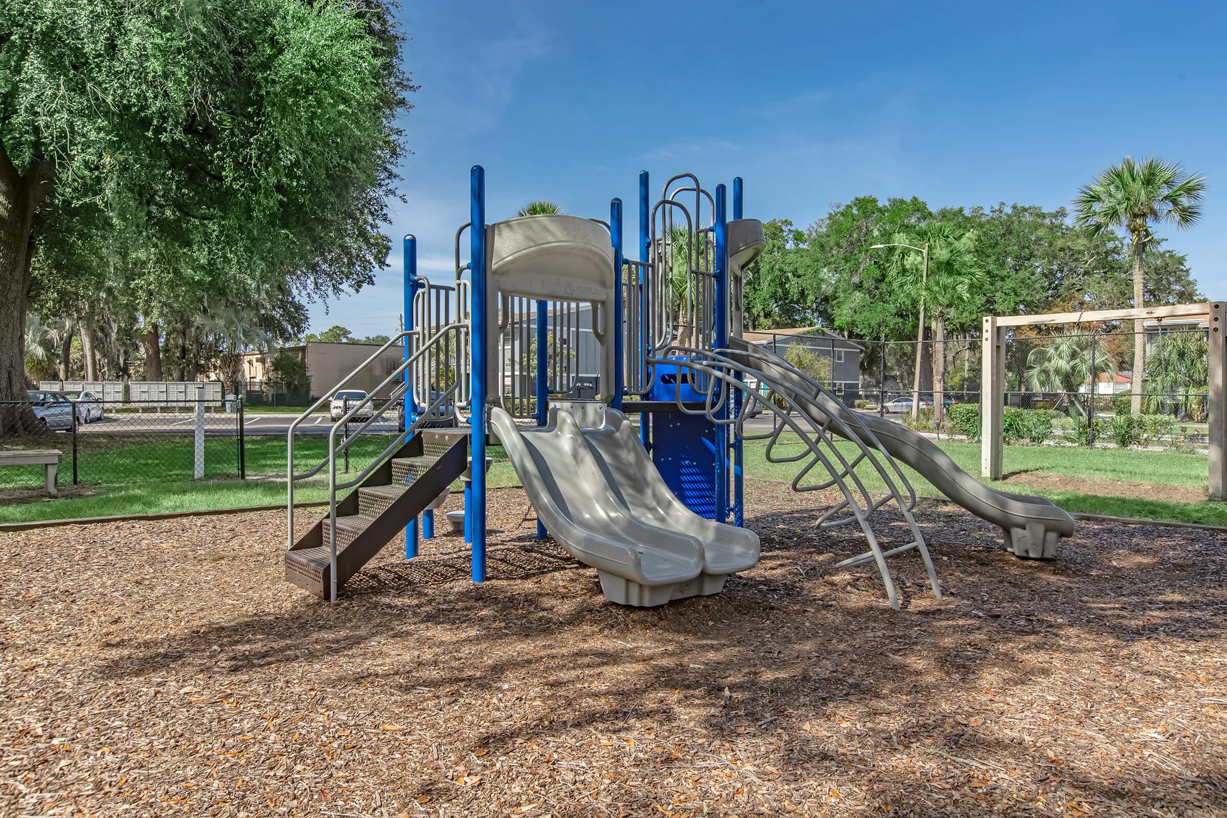 A modern playground featuring two slides, climbing structures, and steps, surrounded by grass and trees. The area is enclosed by a fence, with a clear blue sky in the background, creating a bright and inviting play space for children.