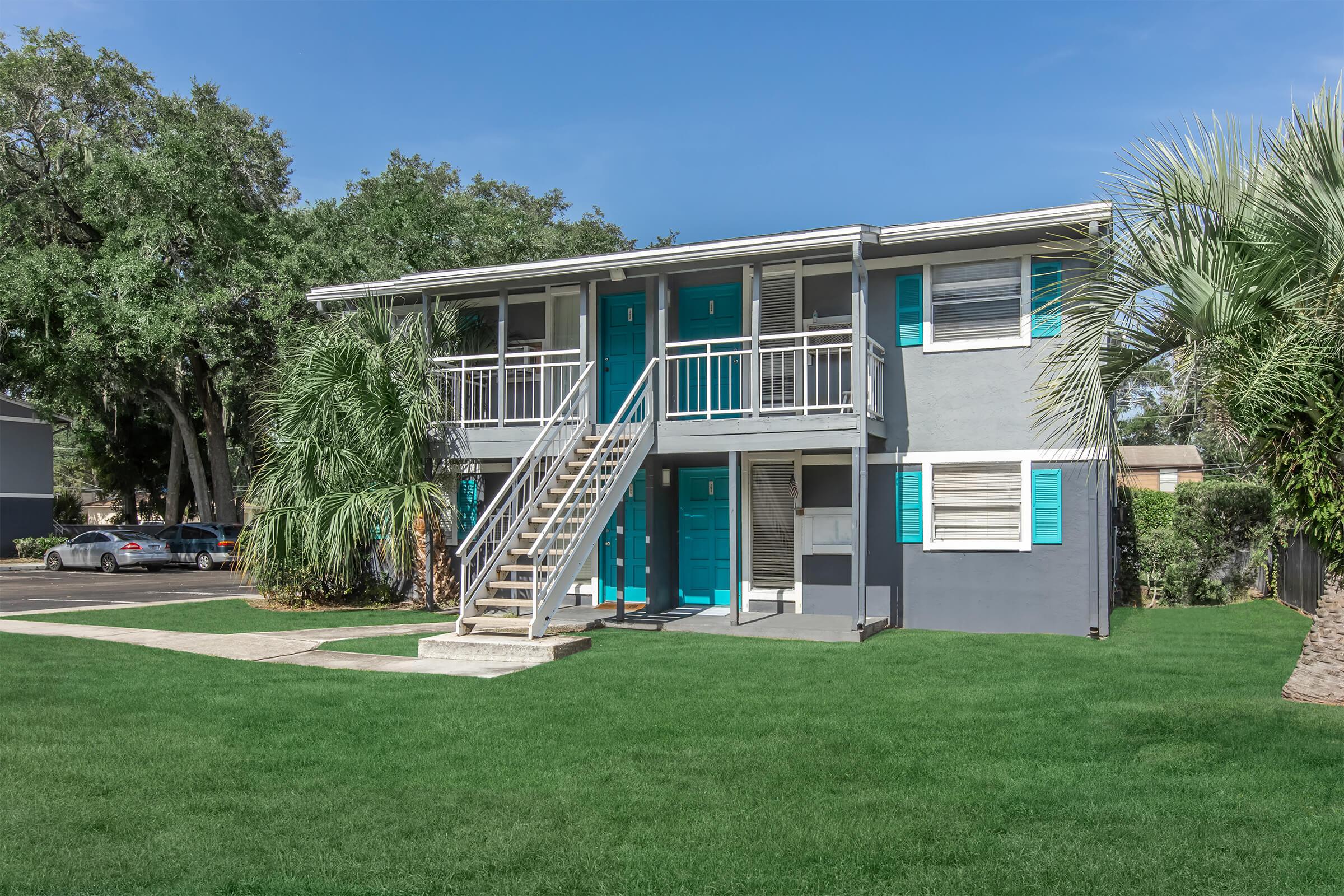 Two-story apartment building featuring a grey exterior with teal doors and shutters, surrounded by lush green grass and palm trees. A concrete pathway leads to the entrance, and cars are parked nearby under a clear blue sky.