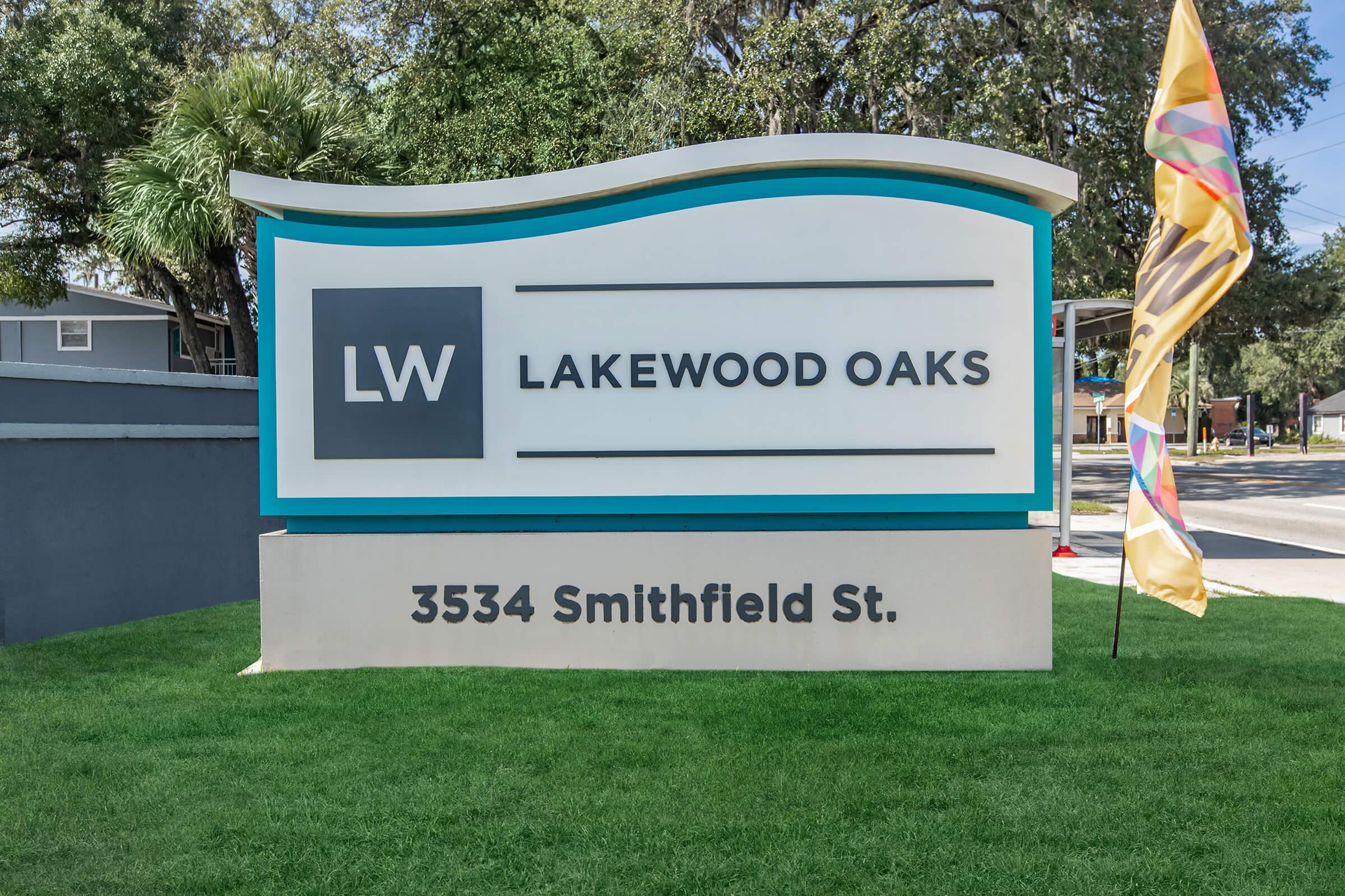 Sign for Lakewood Oaks, displaying the logo "LW" and the address "3534 Smithfield St." The sign features a modern design with a blue and white color scheme, set in a grassy area. A colorful flag is positioned nearby.