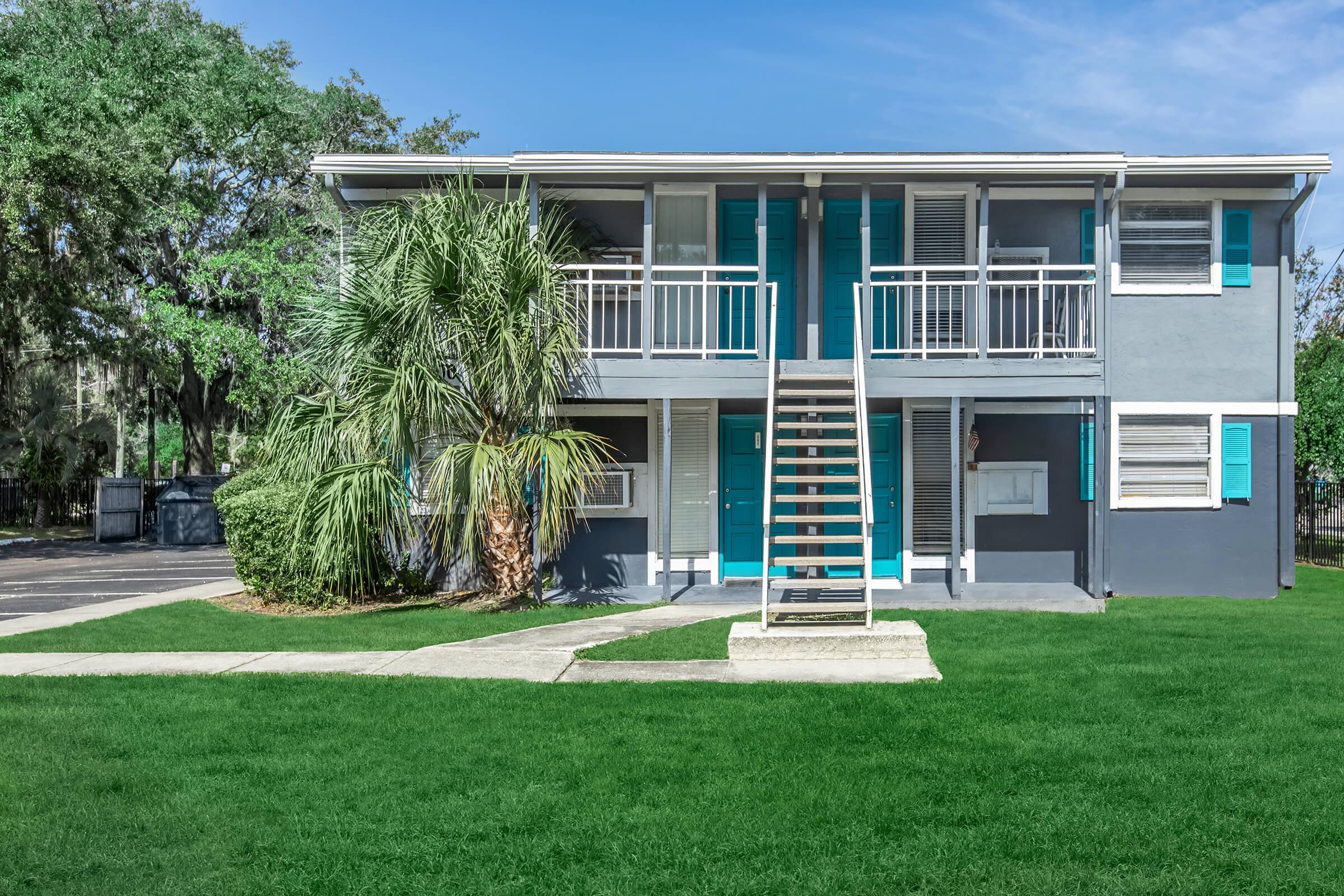 A modern two-story apartment building with blue shutters, set in a grassy area. The building features multiple balconies, an exterior staircase, and palm trees nearby, under a clear blue sky.