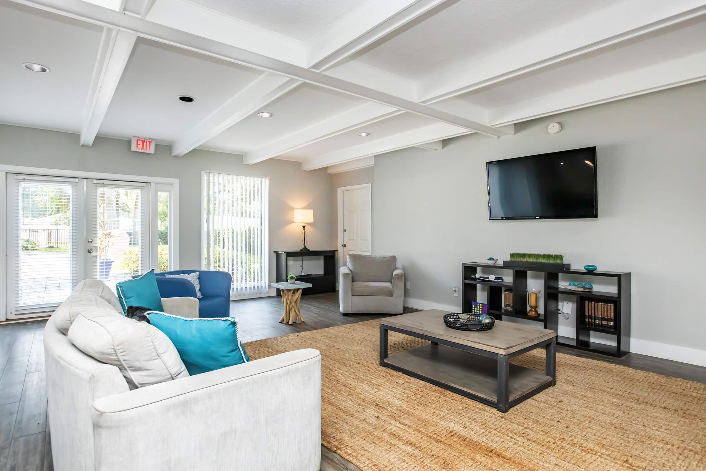 A modern living room featuring a light gray color scheme. It includes a comfortable sofa with teal pillows, a coffee table on a natural fiber rug, and a minimalist TV stand. Large windows allow natural light, with a door leading outside. A cozy armchair is also present, creating a welcoming atmosphere.