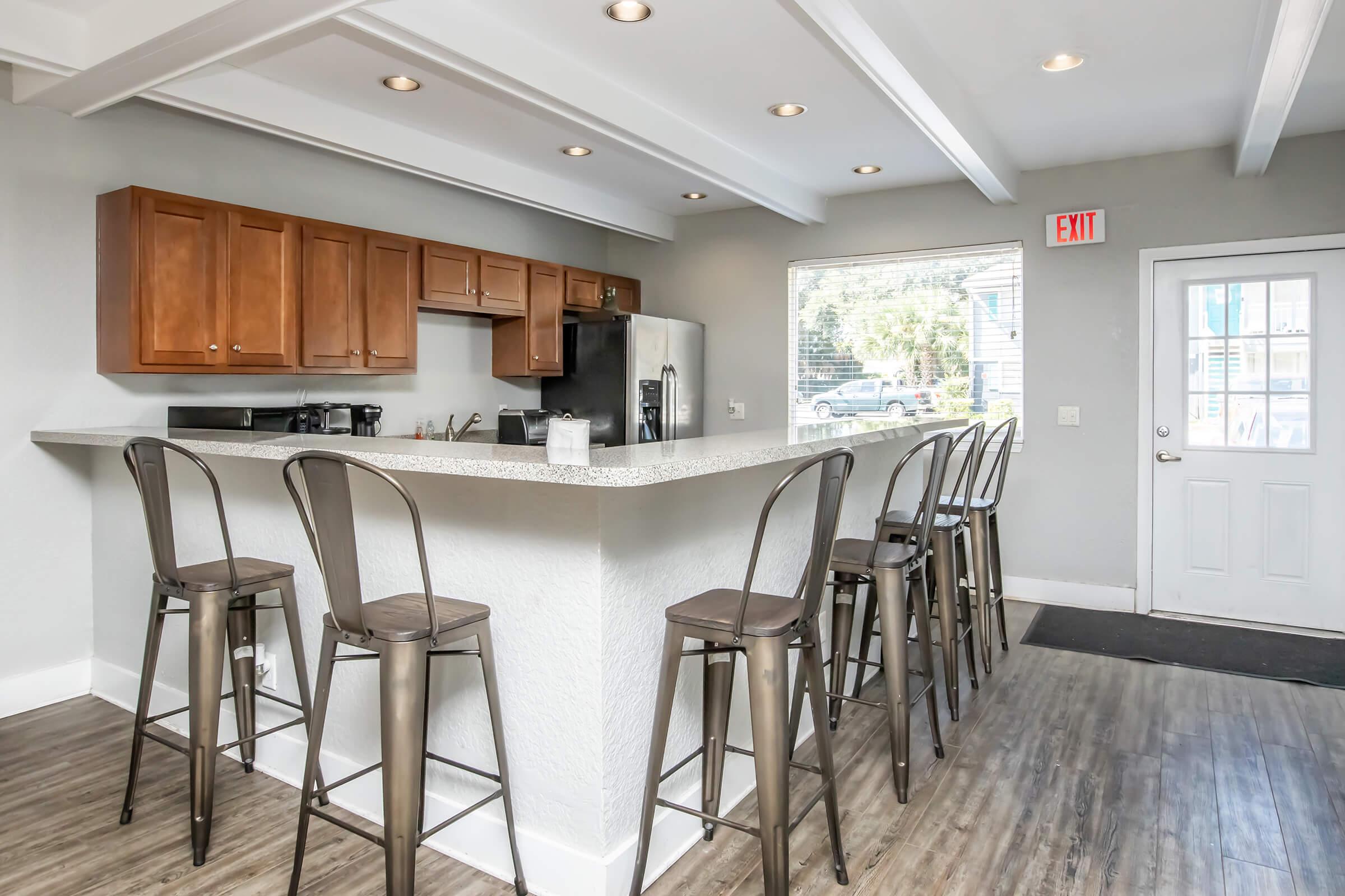 A modern kitchen with a large island bar featuring high metal stools. The cabinetry is wooden, and there are kitchen appliances visible. A door leads outside, and a large window lets in natural light, illuminating the clean and bright interior. The flooring is light wood, enhancing the contemporary look.