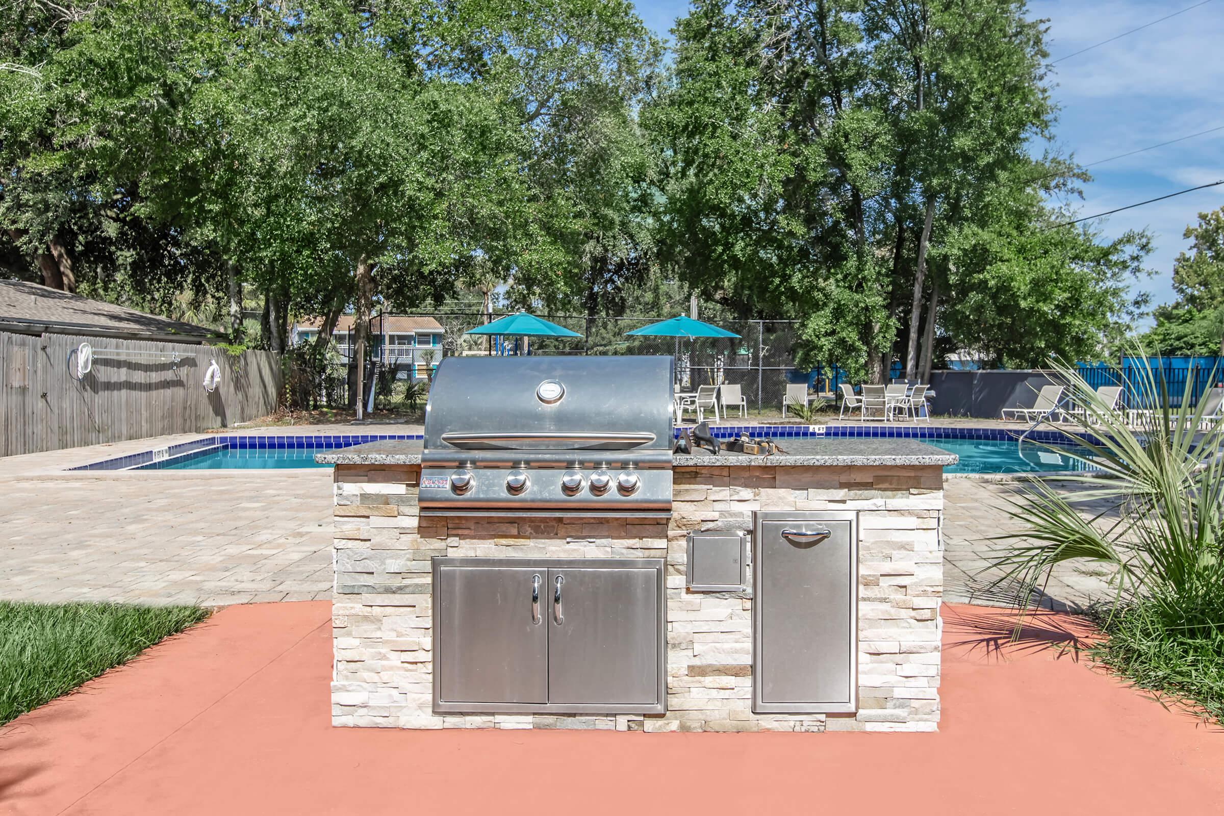A modern outdoor kitchen featuring a stainless steel grill on a stone countertop, set by a swimming pool. The area is surrounded by lush trees, lounge chairs, and umbrellas, creating a relaxing environment for outdoor dining and entertainment.