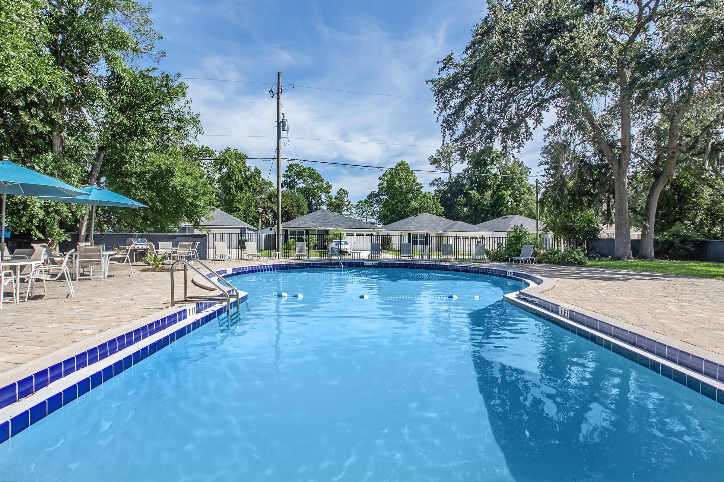 A serene swimming pool surrounded by trees and residential buildings. The pool features a clean, blue surface with lounge chairs and umbrellas on the deck, inviting for relaxation. Clear skies and gentle lighting enhance the peaceful atmosphere of the area.
