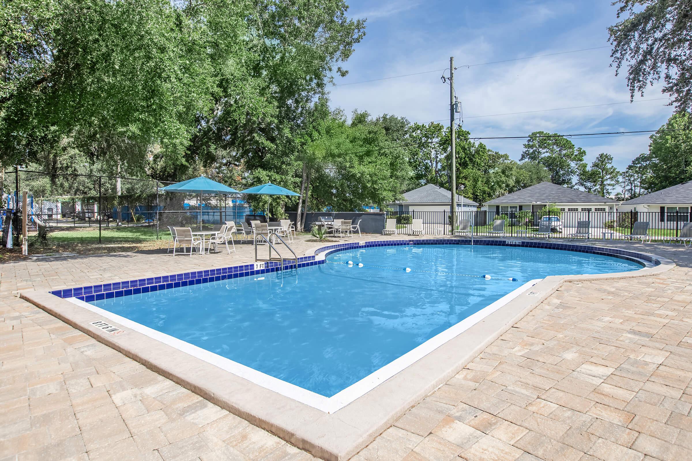 A clear blue swimming pool surrounded by a spacious patio made of stone pavers. There are lounge chairs and umbrellas nearby, along with grassy areas and trees in the background. The sky is partly cloudy, enhancing the inviting atmosphere of the outdoor space.