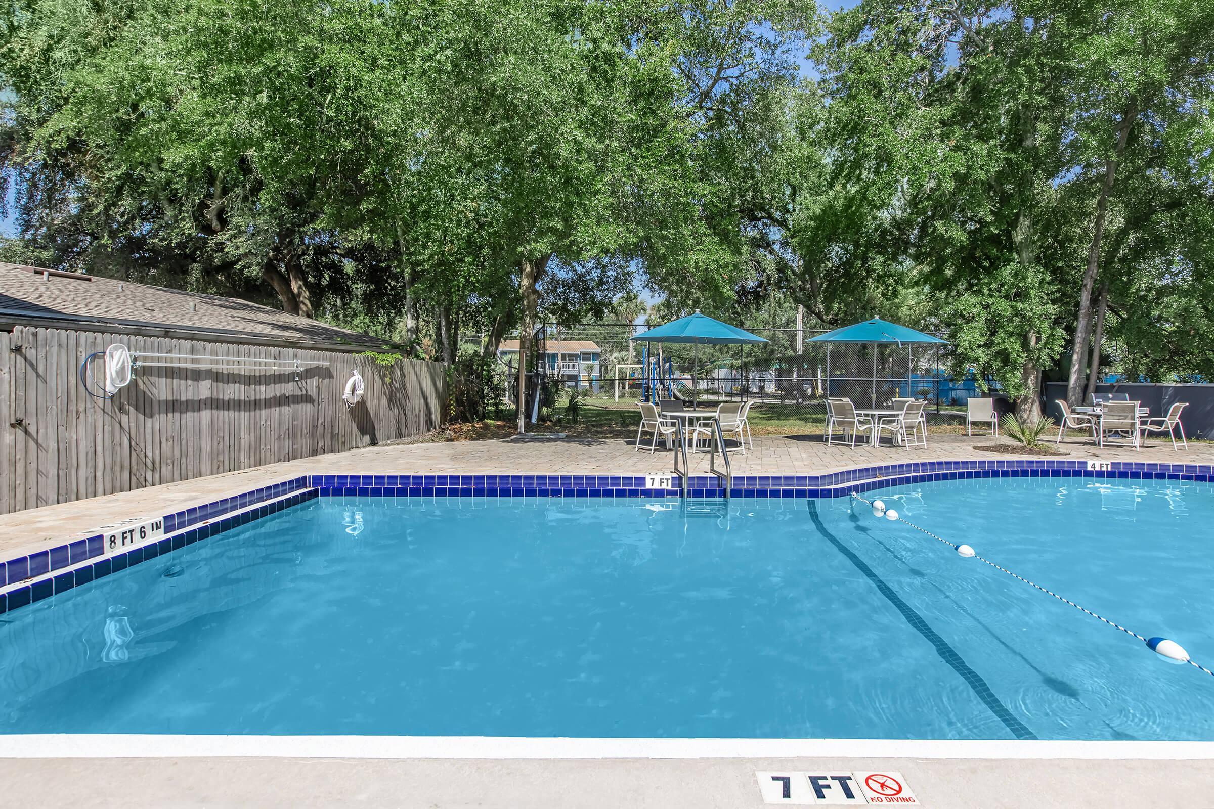 A swimming pool surrounded by trees, featuring clear blue water, a shaded area with green umbrellas, and lounge chairs. The pool has marked depth indicators, with a shallow end at 1 foot. In the background, there are more seating options and a fenced area.