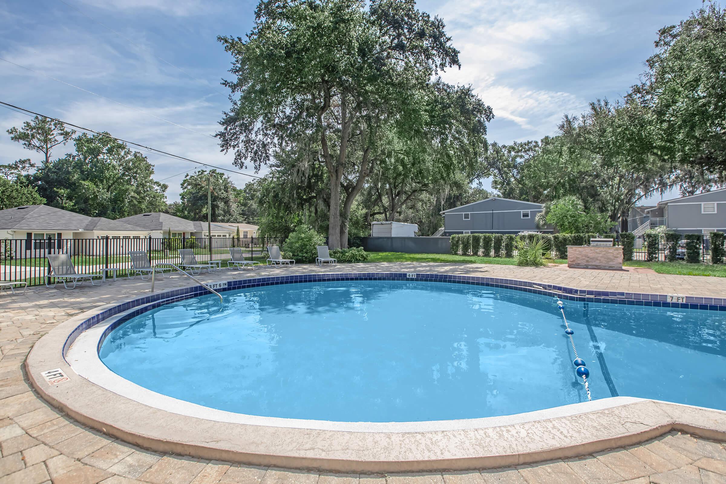 A beautiful, clear blue swimming pool surrounded by patio stones, with lounge chairs on one side. Lush green trees provide shade nearby, and there are houses in the background. A fenced area adds privacy, creating a tranquil outdoor space perfect for relaxation and leisure.