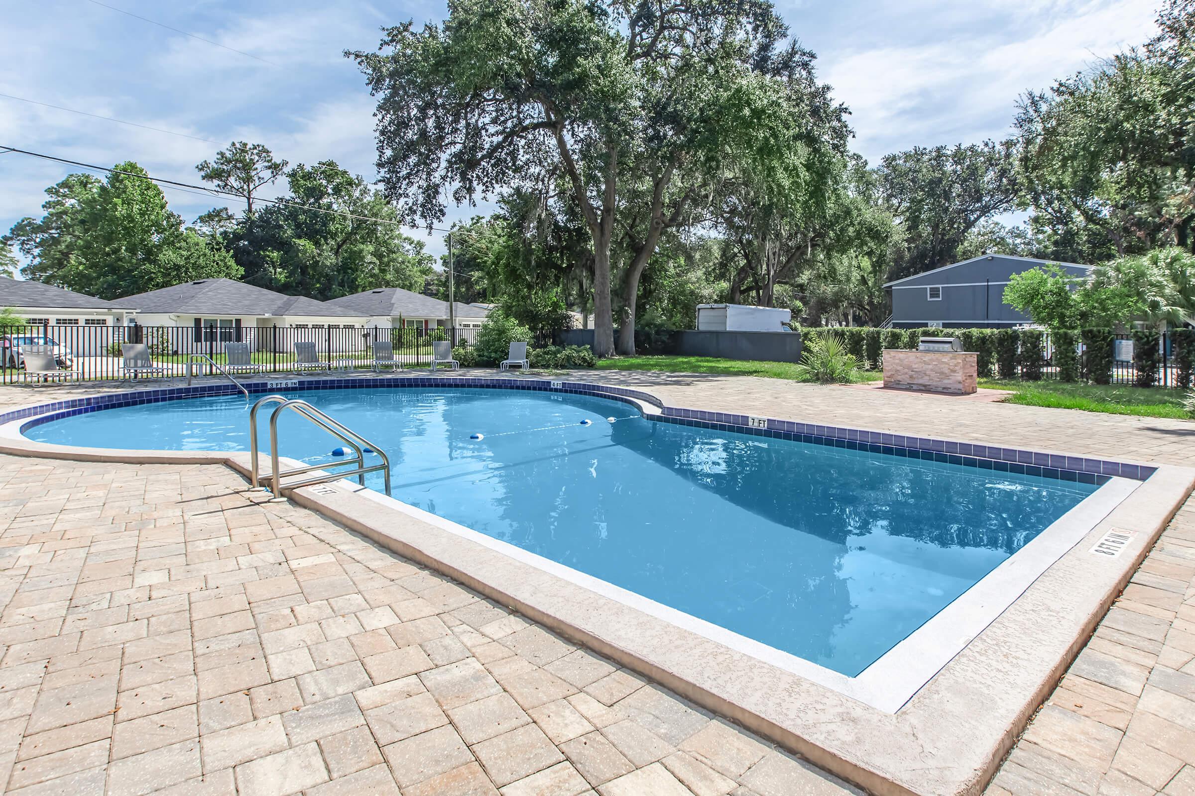 A clear blue swimming pool surrounded by a stone patio. Lush green trees and grass are visible in the background, with a few buildings and a fence nearby. The sunny day creates a bright atmosphere, highlighting the inviting water and outdoor space.