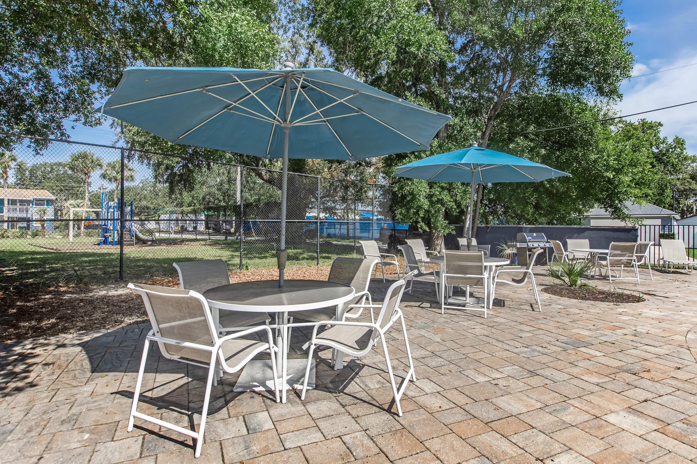 Outdoor patio area with several tables and chairs under large blue umbrellas. The space is surrounded by trees and a fence, with a playground visible in the background. The ground is covered in pavers, creating a welcoming atmosphere for relaxation or outdoor dining.