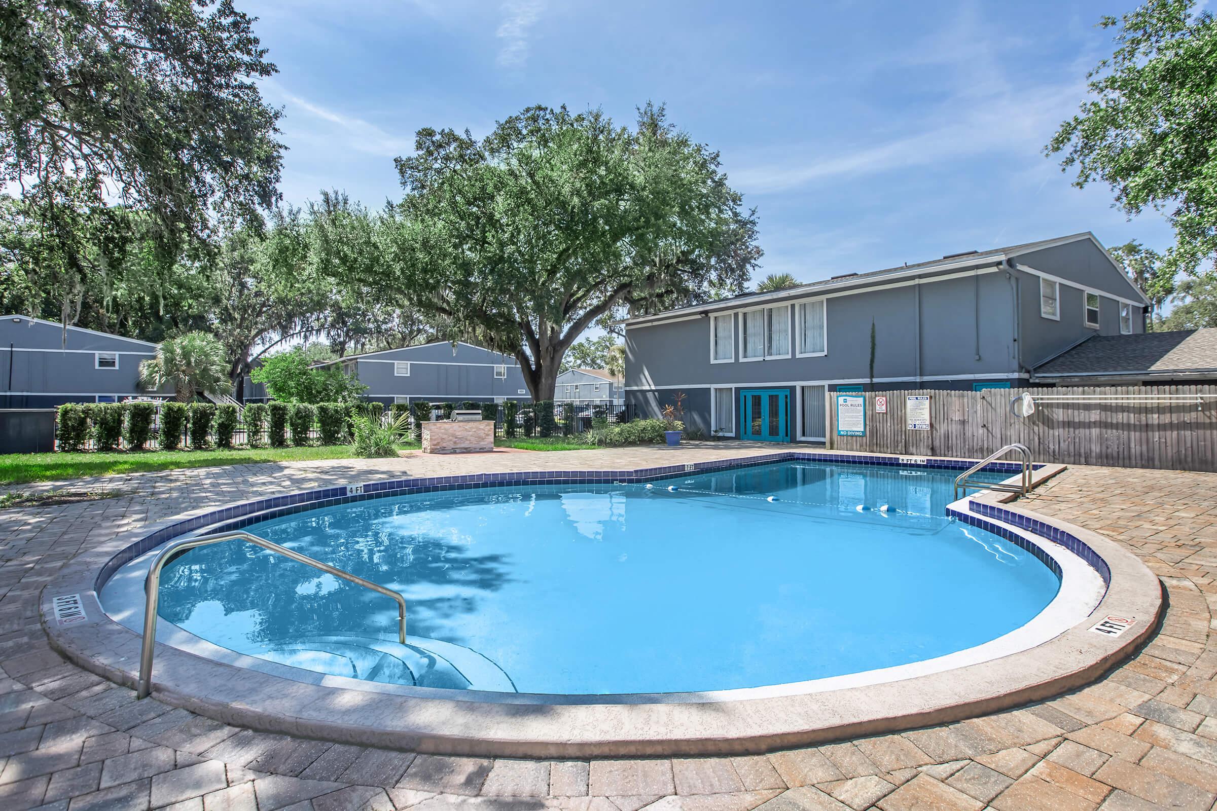 Swimming pool surrounded by a paved deck, featuring a handrail. Lush greenery and trees are in the background, with a two-story gray building on the right and well-maintained landscaping. The pool area is bright and inviting, ideal for relaxation.