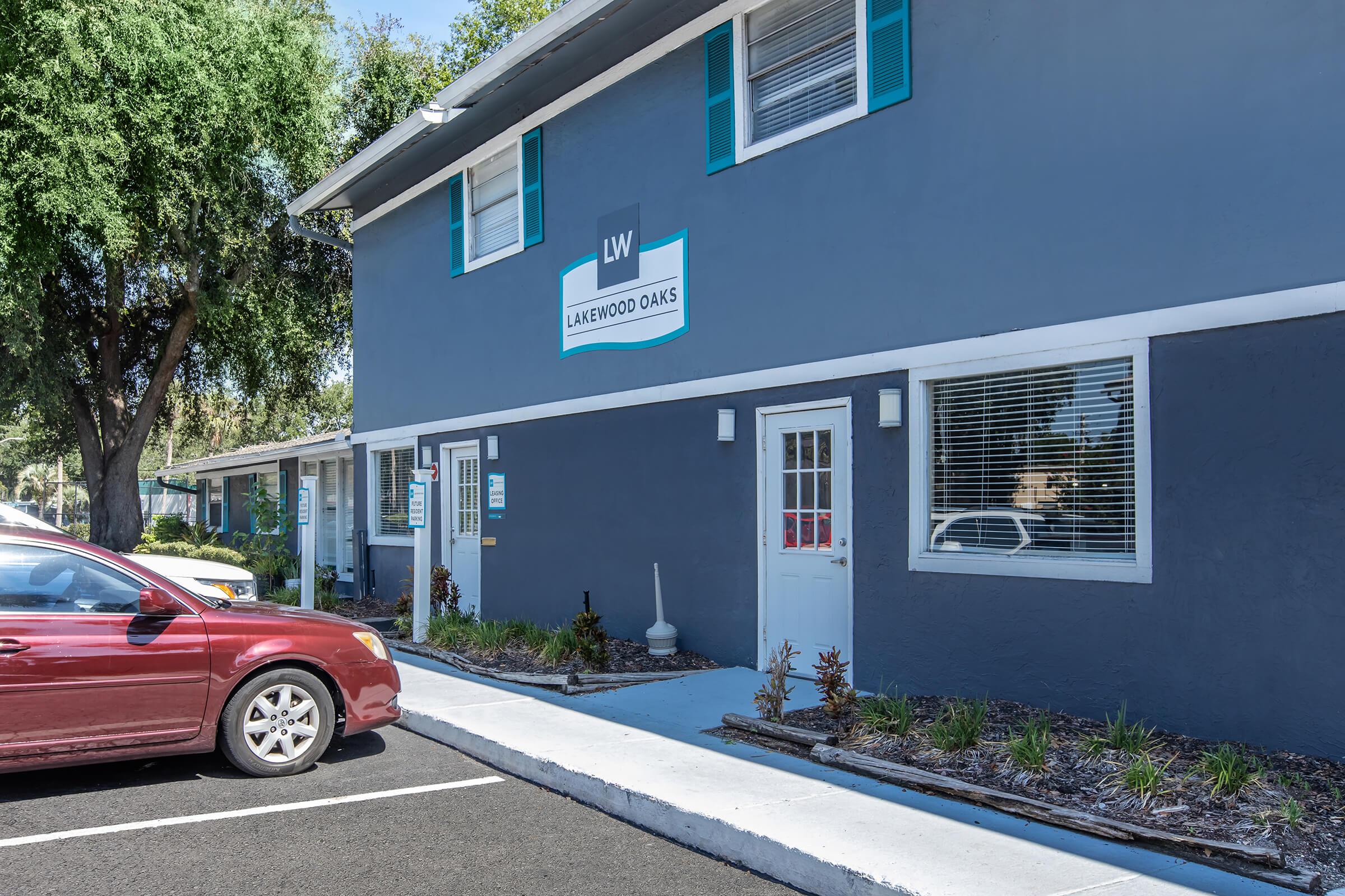 Exterior view of a multi-story building with a blue-gray facade featuring white trim and several windows. The sign reads "LW Lakewood Oaks." In the foreground, a red car is parked alongside a well-maintained walkway with small plants and landscaping.