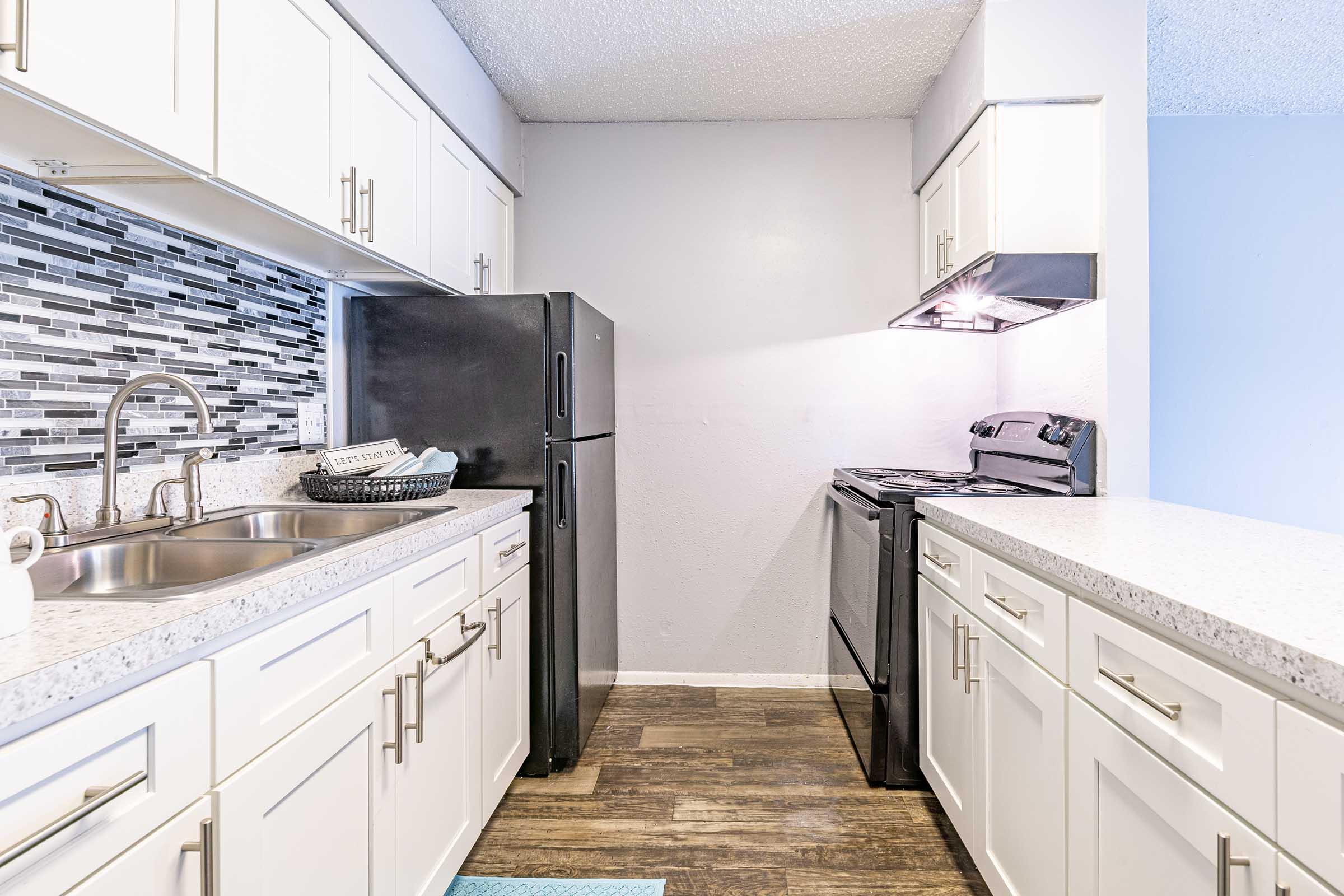 A modern kitchen featuring white cabinets, a black refrigerator, and a black stove. The countertops are light-colored with a speckled design, and a backsplash of gray and black tiles adds visual interest. There’s a double sink, and the flooring is dark laminate, creating a stylish contrast.