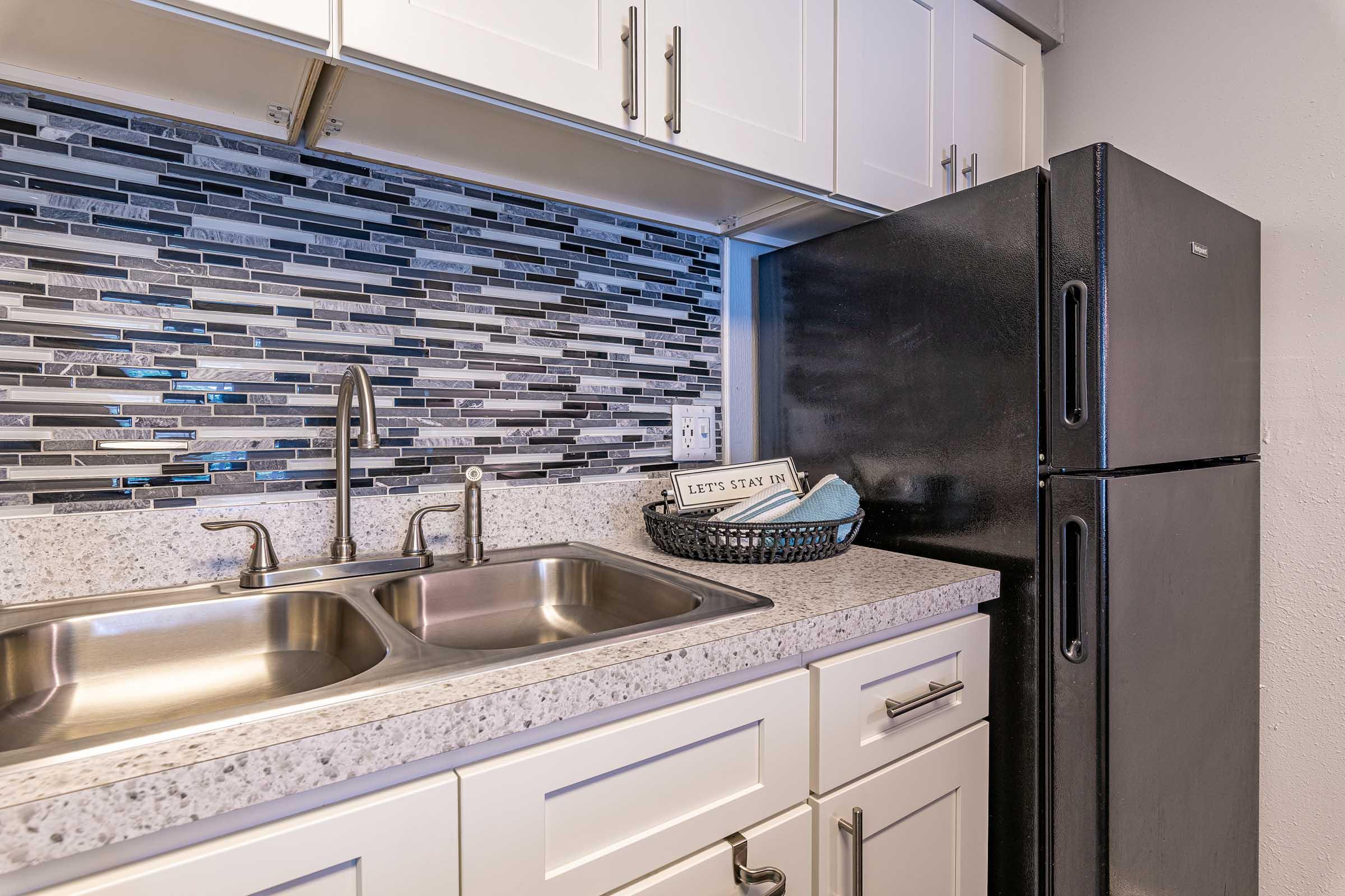 A modern kitchen scene featuring a granite countertop with a double sink, sleek cabinetry, and a black refrigerator. The backsplash is adorned with a mosaic of gray and black tiles, complementing the contemporary design. A decorative basket with text reading "LET'S STAY IN" sits on the counter.