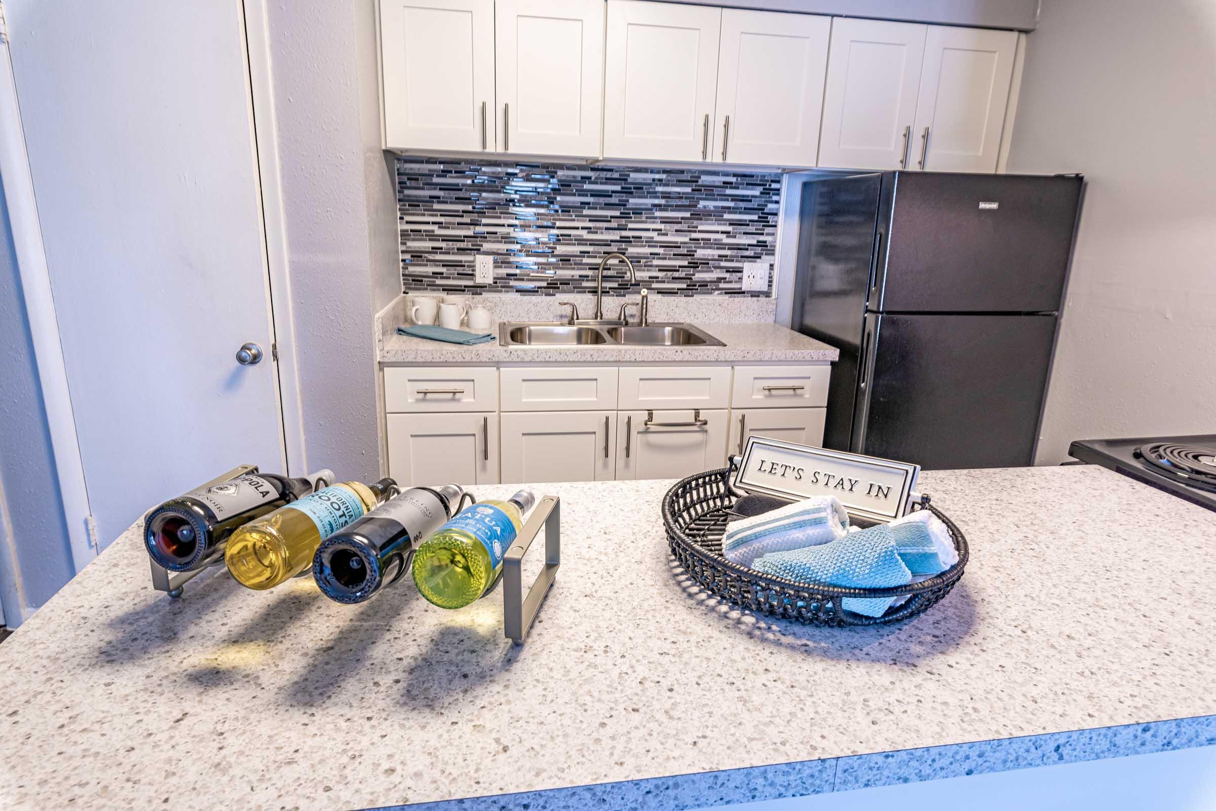A modern kitchen featuring a granite countertop with a wine rack holding several bottles of wine. Behind it is a sleek backsplash with gray tiles and a stainless-steel sink. A black refrigerator is visible, along with a decorative sign that reads "LET'S STAY IN" in a stylish frame. Clean and well-organized, the kitchen exudes a cozy ambiance.