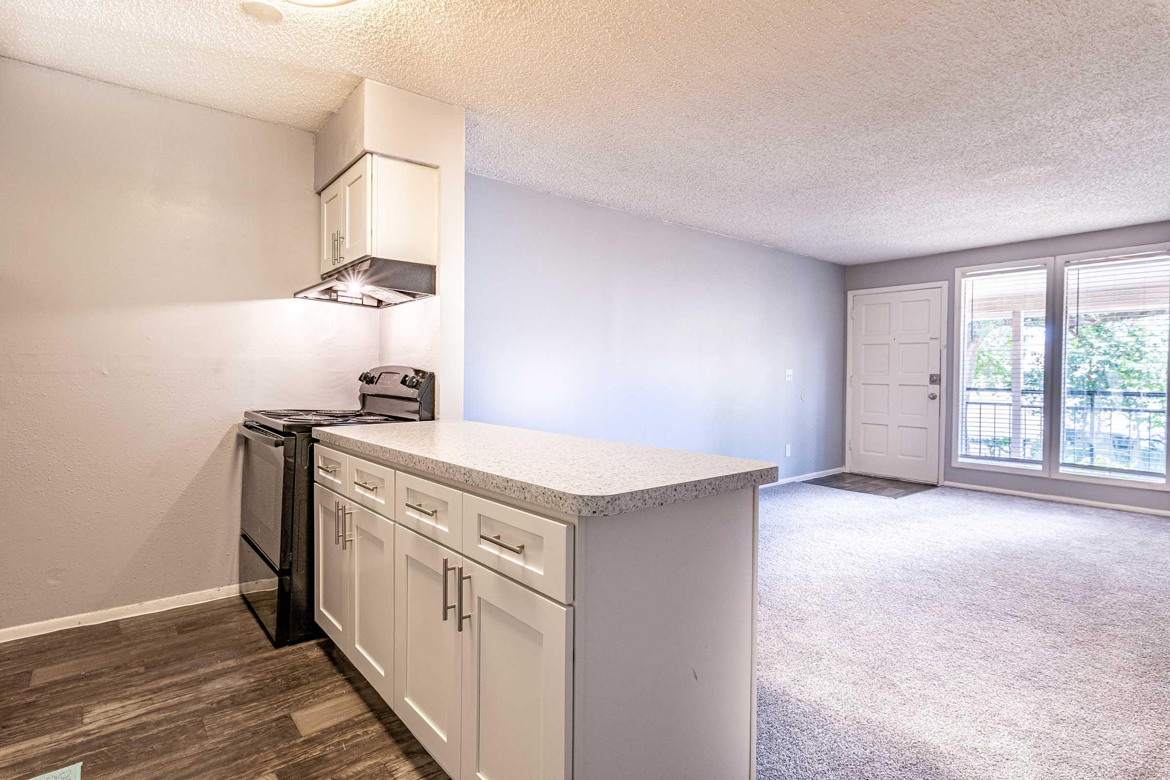 A modern kitchen area featuring white cabinetry and a granite countertop, complete with an oven and stove. The space transitions into a carpeted living room with large windows allowing natural light, leading to an outdoor balcony. The walls are painted in soft, neutral tones.