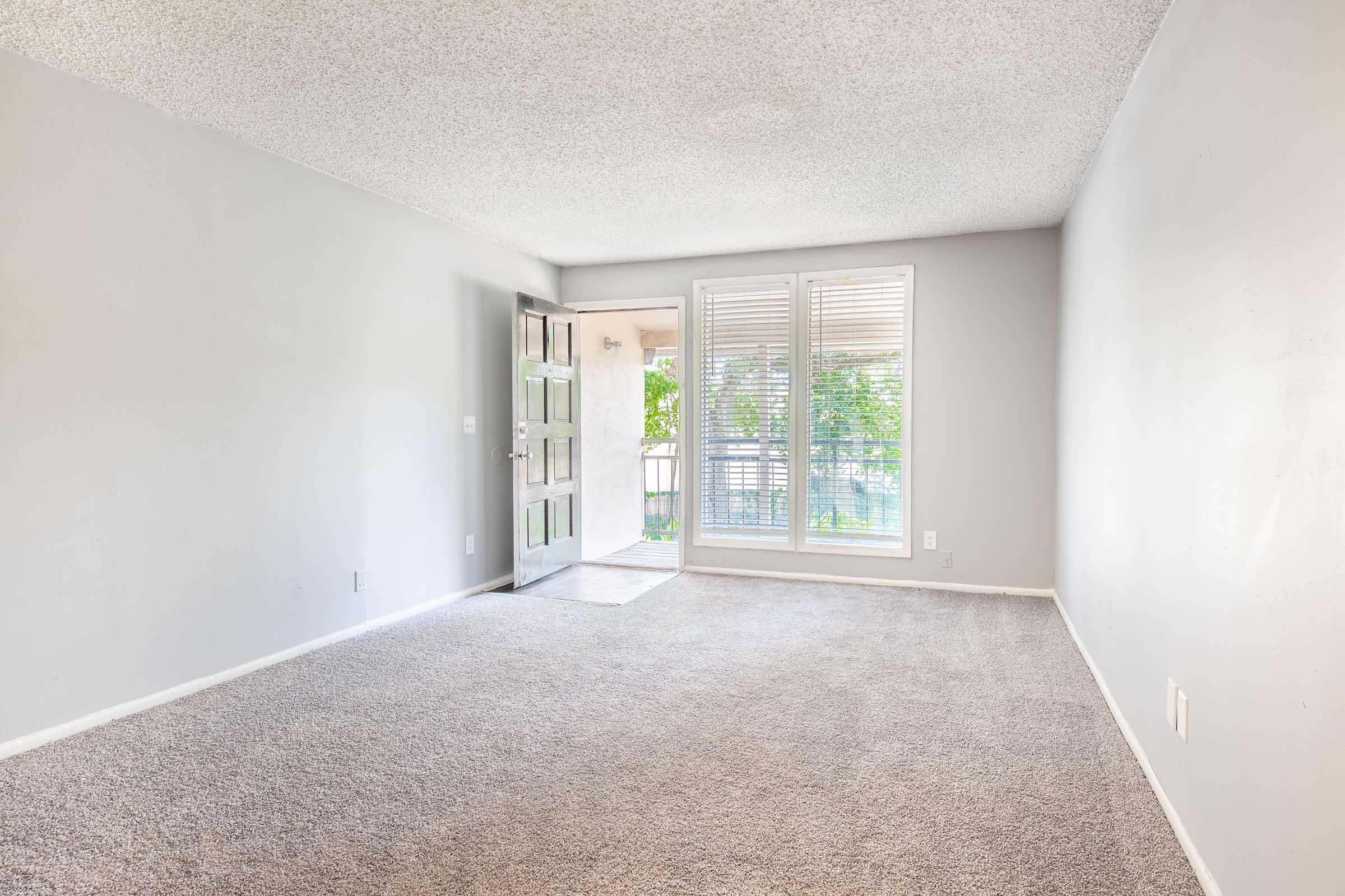 A spacious, empty living room with light gray walls and plush carpet. A large, glass-paneled door leads to a balcony with green trees visible outside. The room features natural lighting from the windows, creating a bright and airy atmosphere.