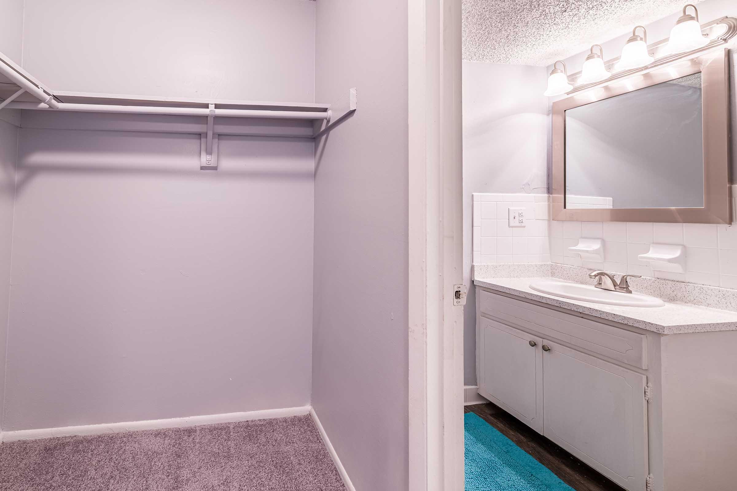 Interior view of a bathroom featuring a vanity with a sink and mirror, illuminated by wall-mounted light fixtures. Adjacent to the bathroom is an empty closet with a hanging rod. The walls are painted light purple, and the floor is partially covered with a teal bath mat.