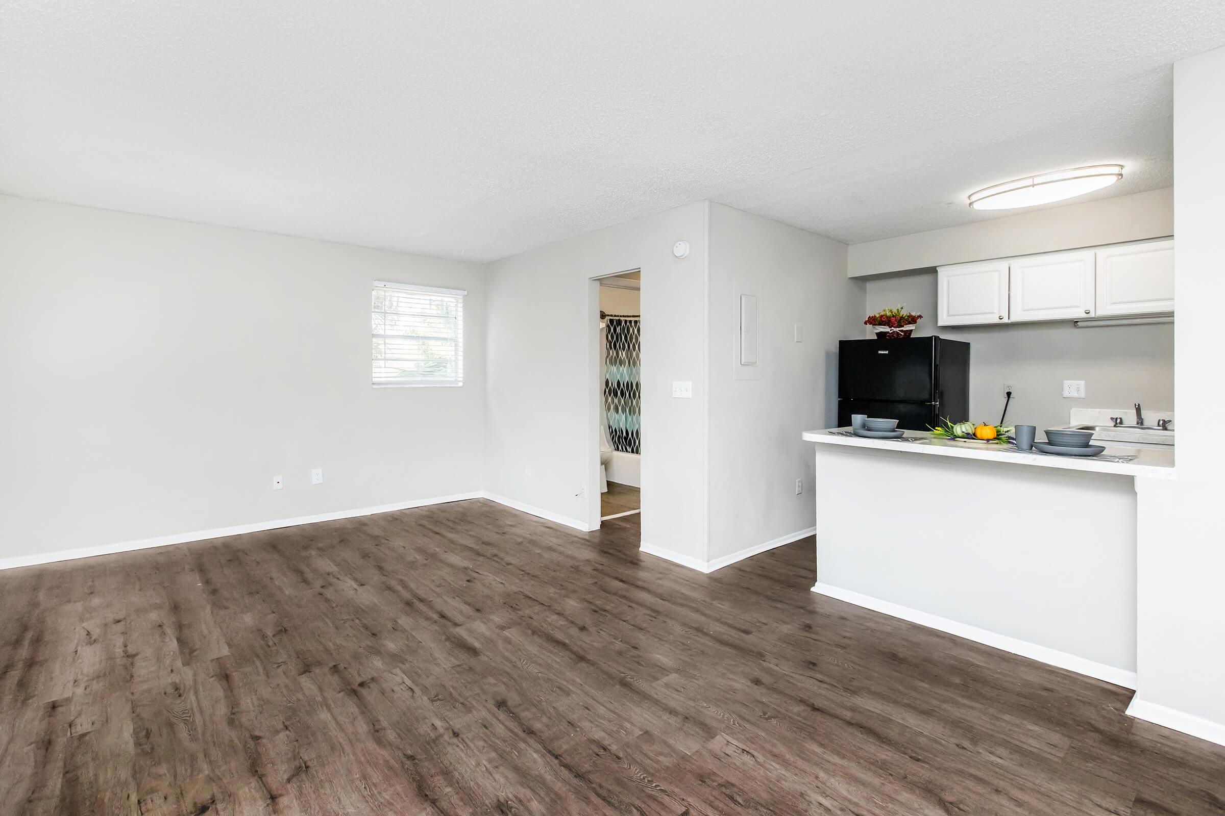 A modern, open-concept living space featuring light gray walls, laminate flooring, and a small kitchen area with a fridge and countertop. There's a window allowing natural light, and a dining area set with plates and a fruit bowl. The overall atmosphere is bright and inviting.
