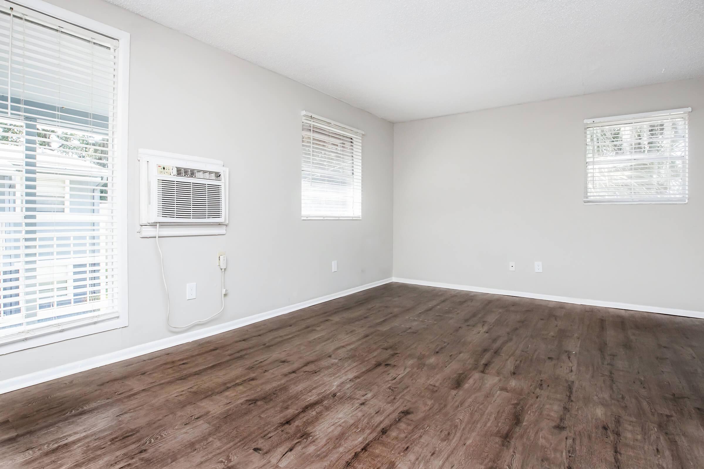 Empty room with light-colored walls and wooden flooring. Two windows on opposite walls allow natural light to enter. An air conditioning unit is mounted on one wall. The space is simple and clean, suitable for various purposes like living or office use.