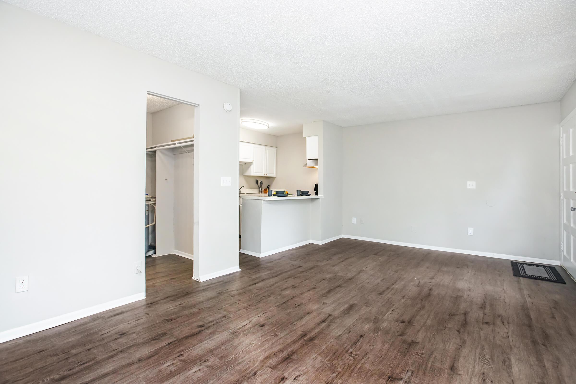 Empty living space with light gray walls and a textured ceiling. The floor is dark laminate wood. To the left, there's an open doorway leading to a closet. In the background, a small kitchen area is visible with white cabinets and appliances. Natural light brightens the room.