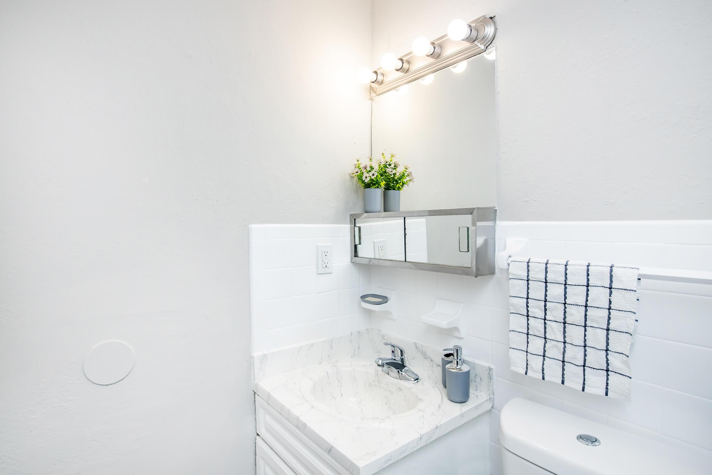 A clean and modern bathroom with a white vanity, a marble countertop, and a mirror with bright round lights above. A small plant sits atop the mirror. There is a neatly folded white towel with blue stripes on a towel rack, and a gray soap dispenser beside the sink. The walls are painted light gray.
