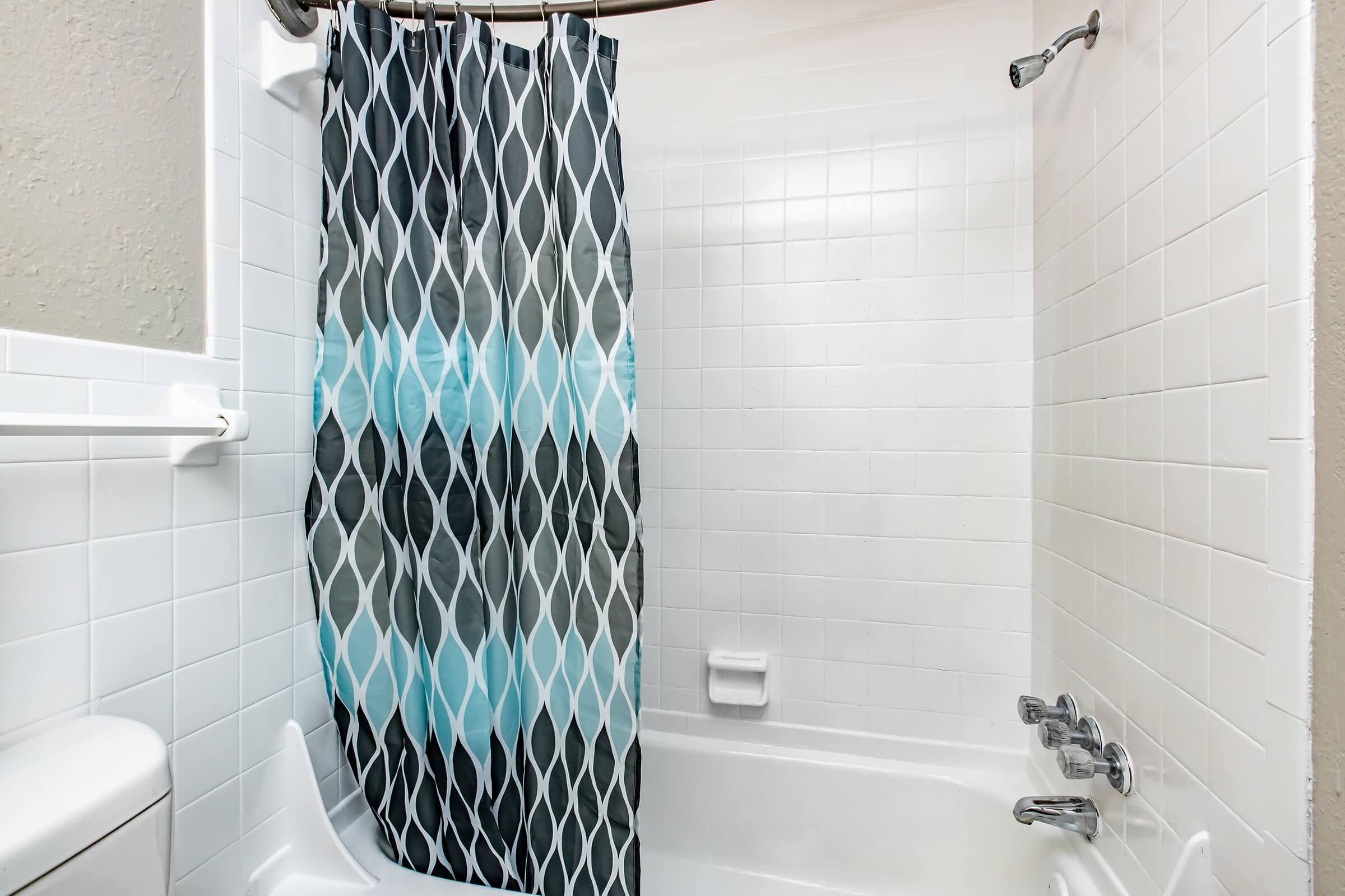 A clean bathroom featuring a white tiled shower with a modern, patterned shower curtain in shades of gray and blue. The space includes a white bathtub, a towel rack, and chrome fixtures.