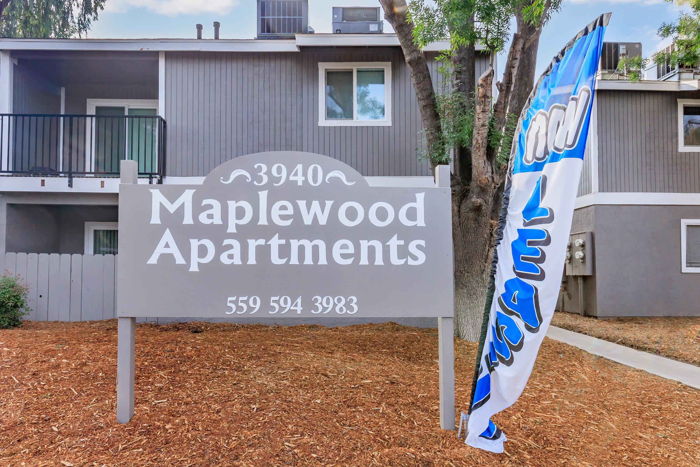 A sign for Maplewood Apartments located at 3940, featuring the phone number 559-594-3983. The sign is situated near a tree and a flag fluttering beside it. The background shows a two-story apartment building with balconies.