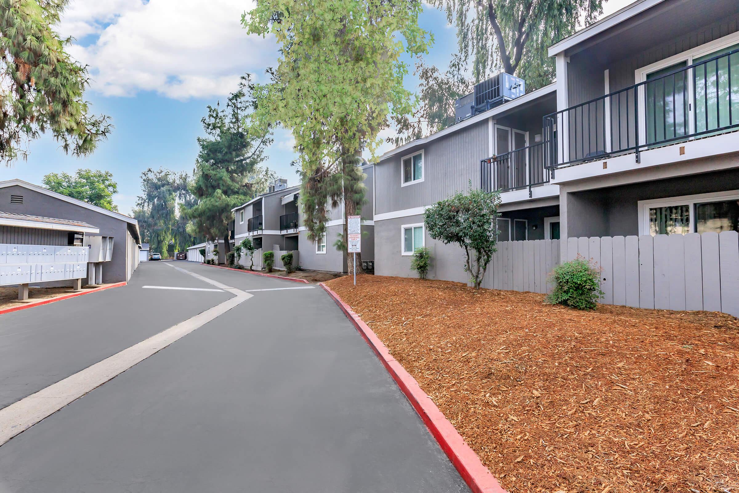 A view of a residential complex featuring a paved road curving through the property, lined with trees and shrubs. The buildings are two stories with balconies, surrounded by freshly mulched landscaping. A clear blue sky is visible, adding to the serene atmosphere.