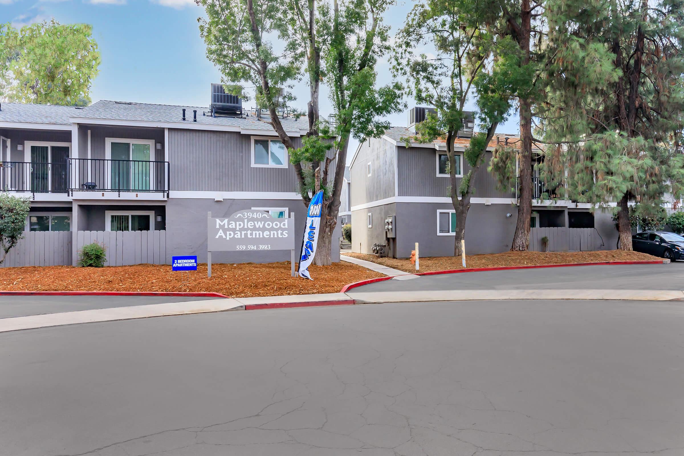 Maplewood Apartments exterior view featuring two-story buildings surrounded by greenery. A sign indicates the apartments' name, and there is a parking area with trees providing shade. The entrance is visible, with a roadway curving around the property.