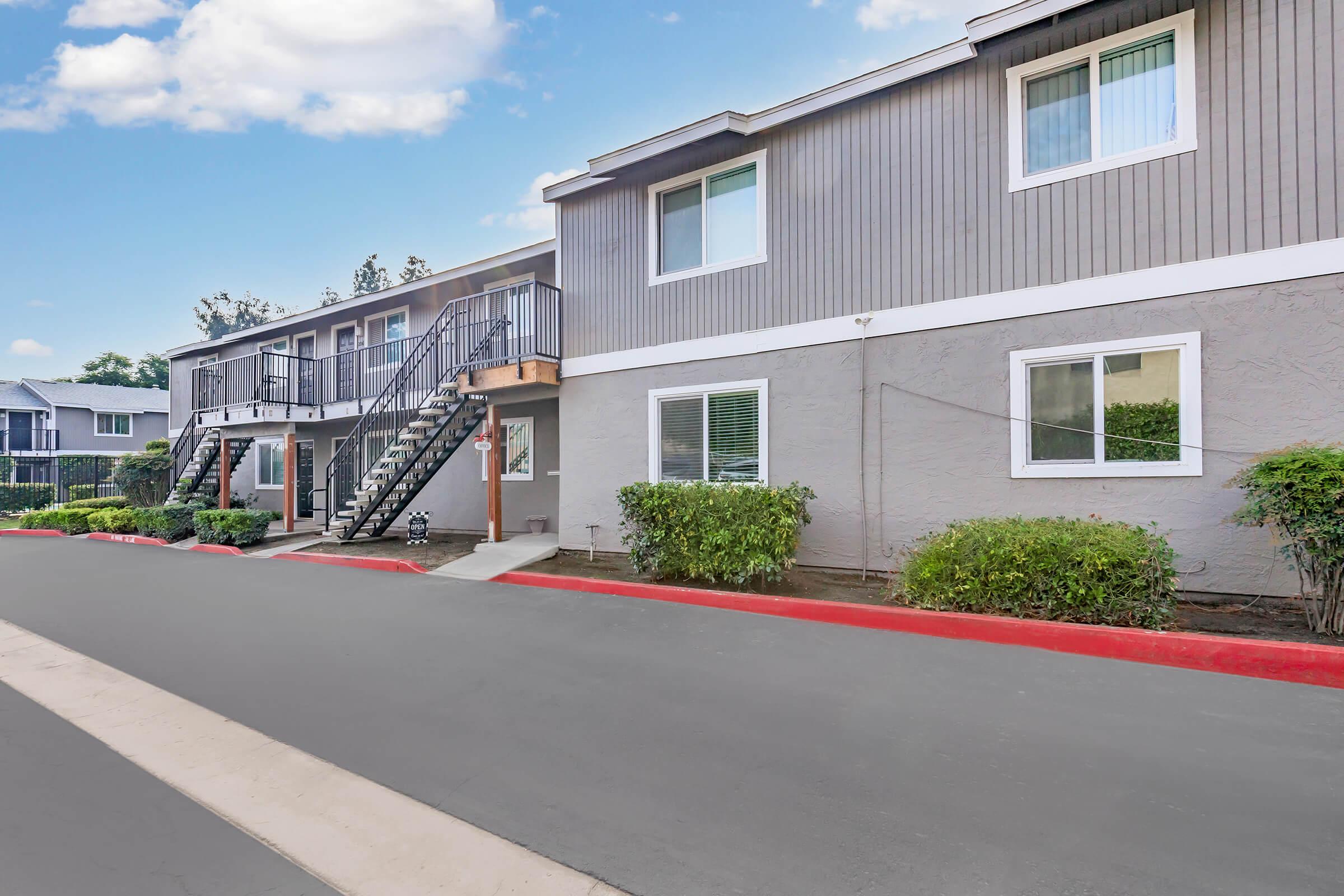 Exterior view of a multi-unit apartment building featuring two levels, stairs leading to the upper units, and greenery along the pathway. The sky is clear with a few clouds, and the pavement is newly painted with a red line along the edge.