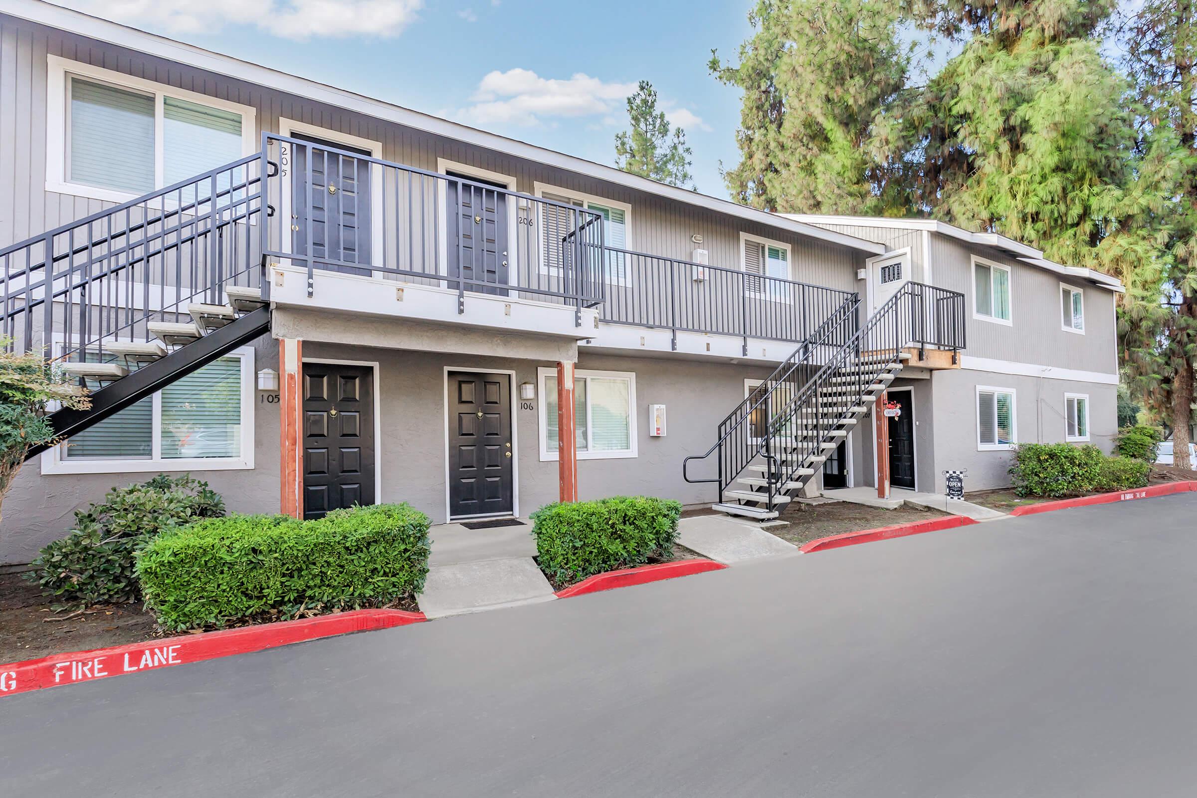 Exterior view of an apartment building with a stairway leading to the upper units. The building has gray siding, dark doors, and landscaped greenery in front. A marked fire lane is visible in the foreground, and trees line the background under a clear blue sky.