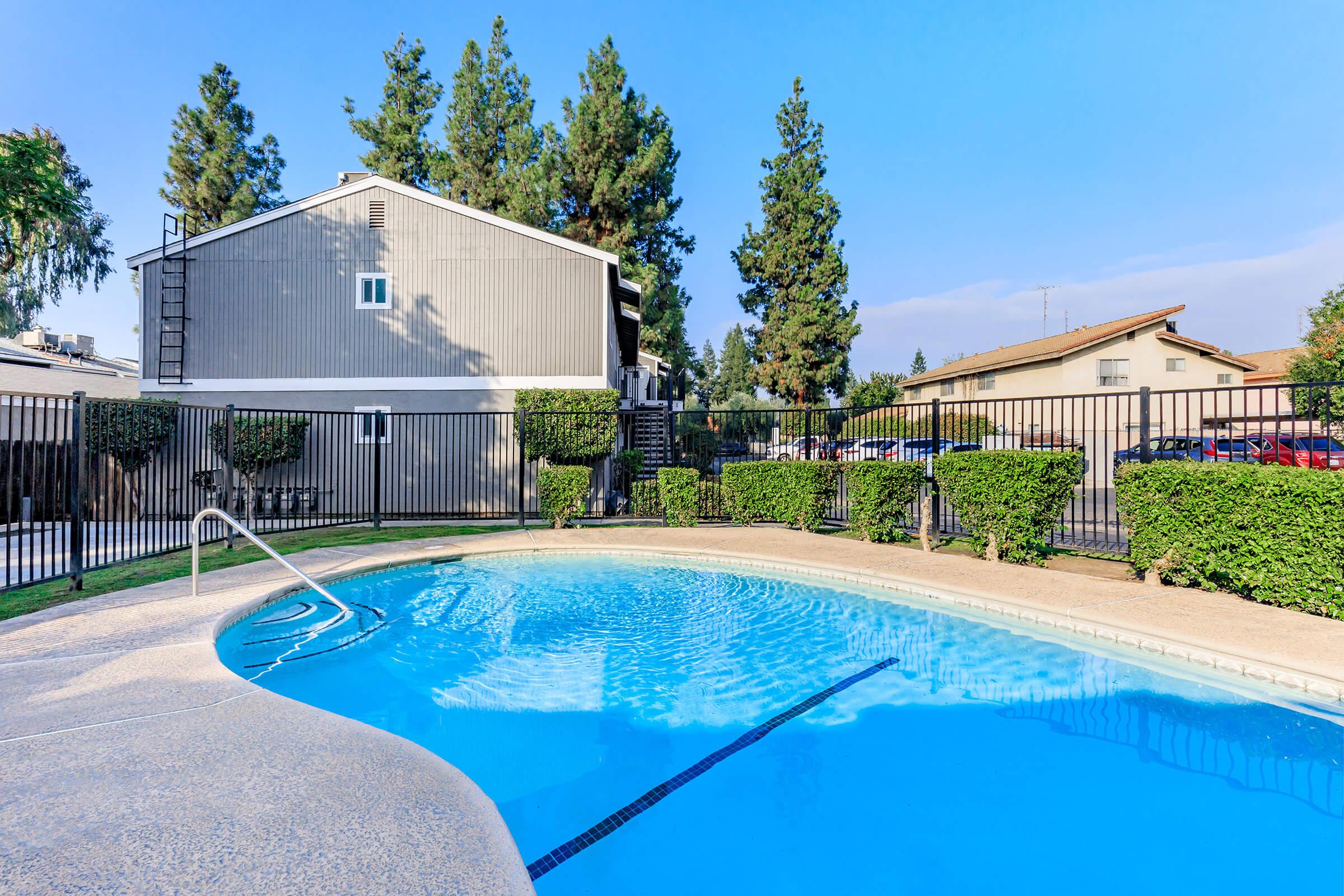 A clear swimming pool surrounded by a fenced area with landscaped hedges. In the background, there are two multi-unit buildings with trees and a blue sky overhead. The pool has a ladder and a gentle curve along its edge, inviting for relaxation and leisure.