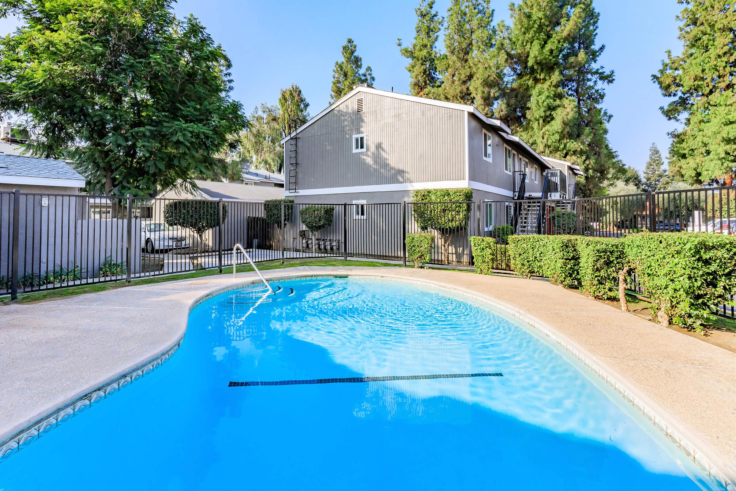 A clear blue swimming pool surrounded by well-manicured bushes and trees. In the background, there are gray apartment buildings and a fenced area. The scene is bright and sunny, indicating a warm day. A ladder leads into the pool, enhancing the inviting atmosphere.