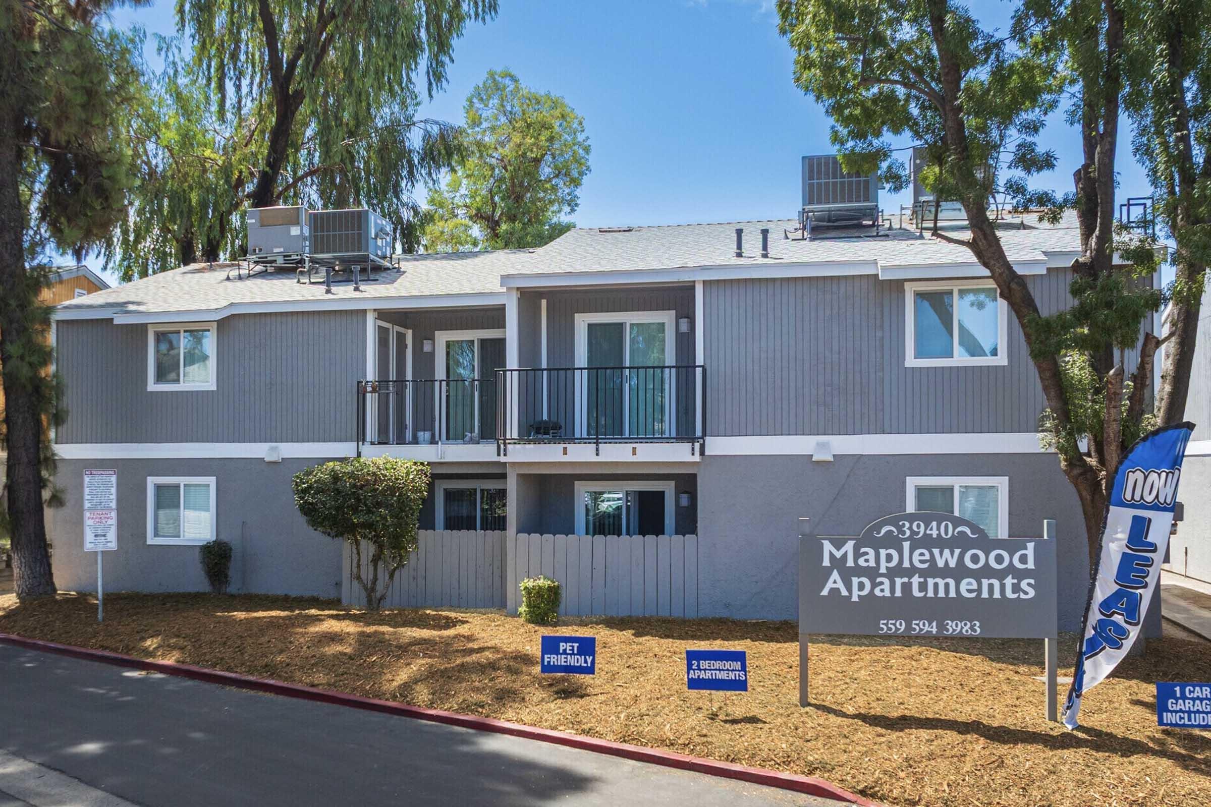 Exterior view of Maplewood Apartments featuring a two-story building with a gray facade, balconies, and a landscaped area. The sign indicates the name and contact information for leasing. There are multiple air conditioning units on the roof, and the surroundings include trees and shrubbery.