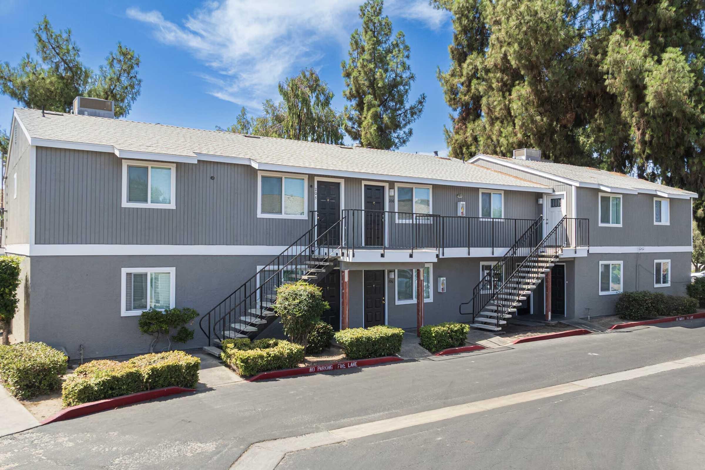 Two-story residential apartment building with a gray exterior, featuring several balcony entrances and adjacent walkways. There are neatly trimmed bushes along the front, with a paved street visible in the foreground and trees in the background under a clear blue sky.