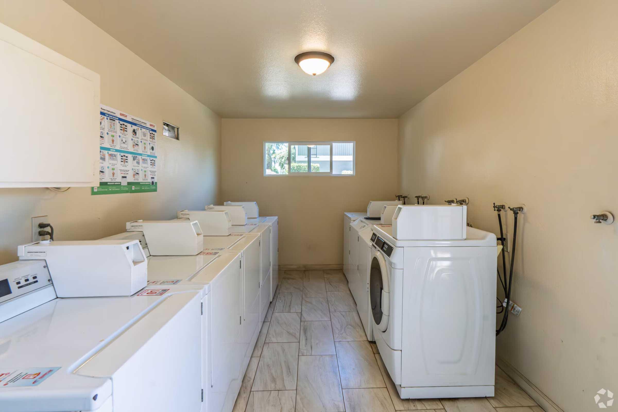 A clean and well-lit laundry room featuring several white washing machines and dryers lined up against the wall. The room has light-colored walls and a tile floor. A window lets in natural light, and laundry information is displayed on a board.