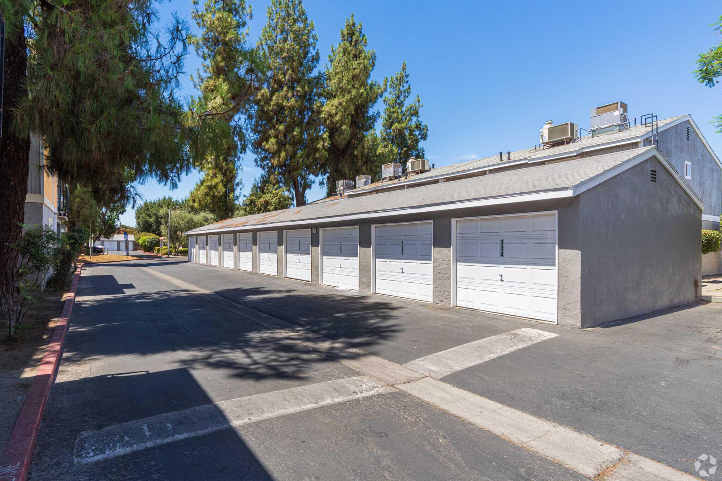 A row of garage doors in a residential area, surrounded by trees. The garages have white doors and are set along a wide asphalt driveway under bright blue sky, indicating a sunny day.