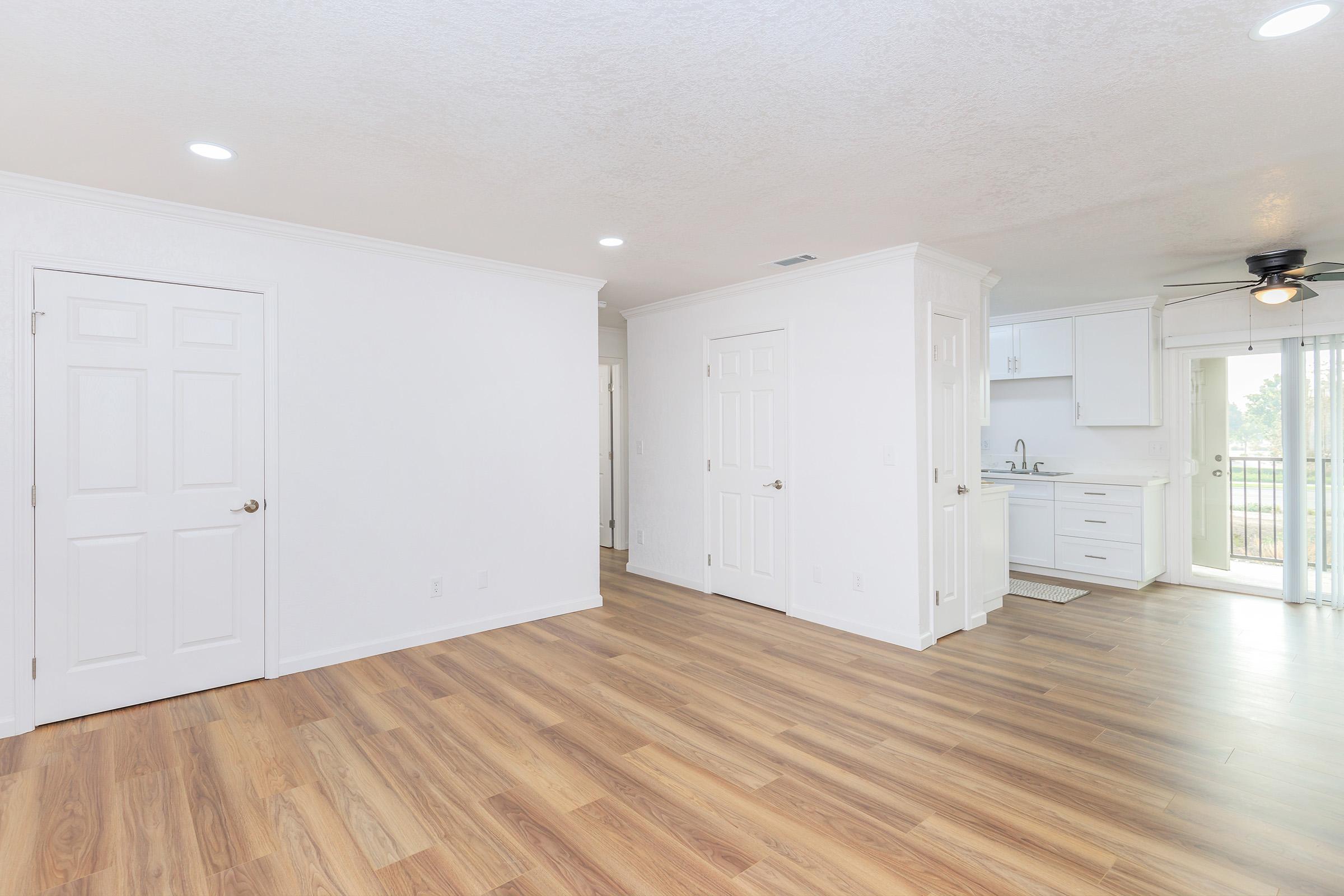 A bright and spacious room featuring hardwood flooring, white walls, and modern fixtures. There is a doorway leading to another room, a kitchenette area with white cabinets, and a sliding glass door that allows natural light and access to an outdoor space.