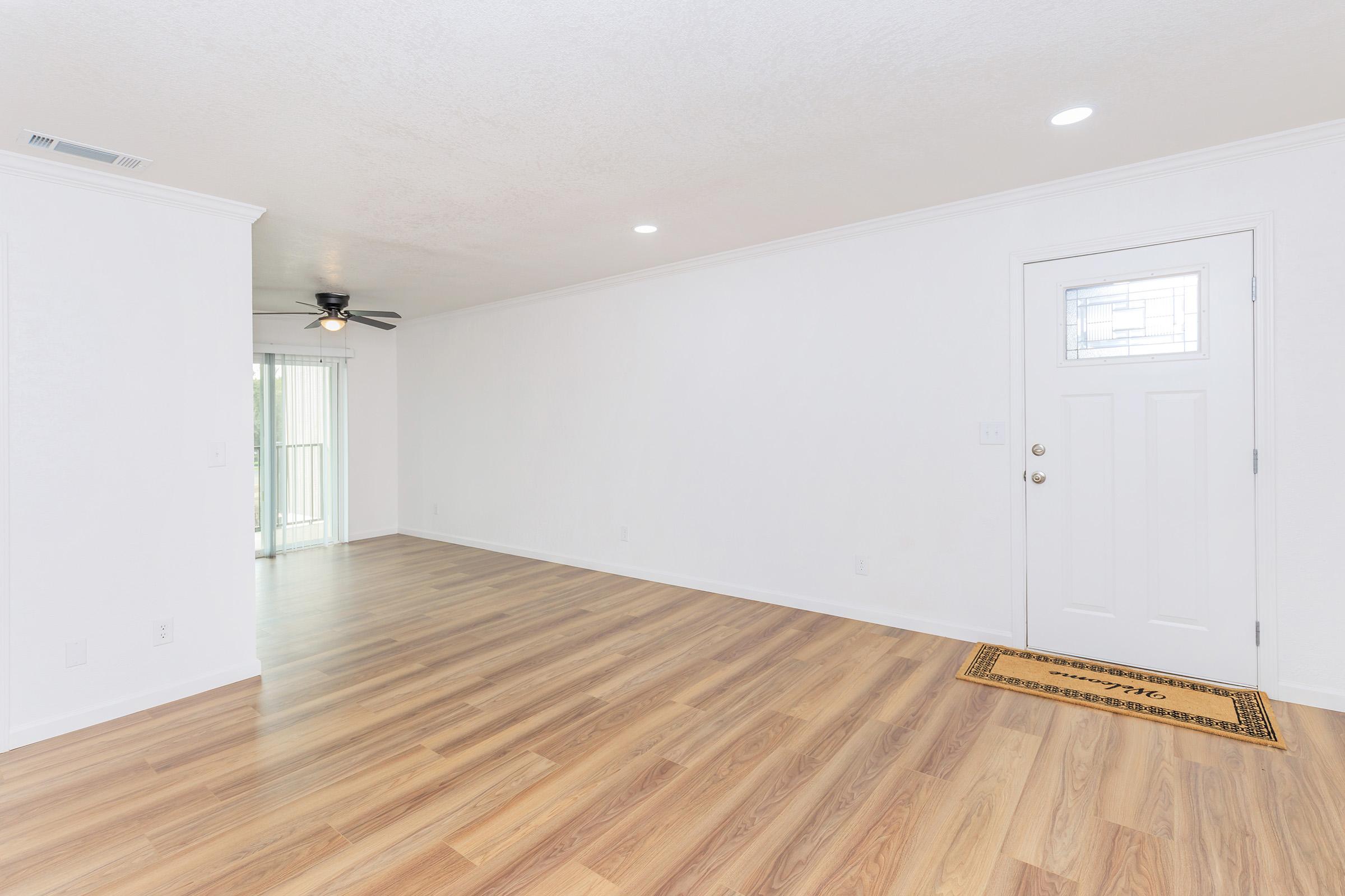 A bright, spacious living room with light-colored walls and polished wooden flooring. There is a ceiling fan, an entrance door with a decorative glass panel, and a welcome mat by the door. Natural light comes in from a sliding door leading to an outdoor space.