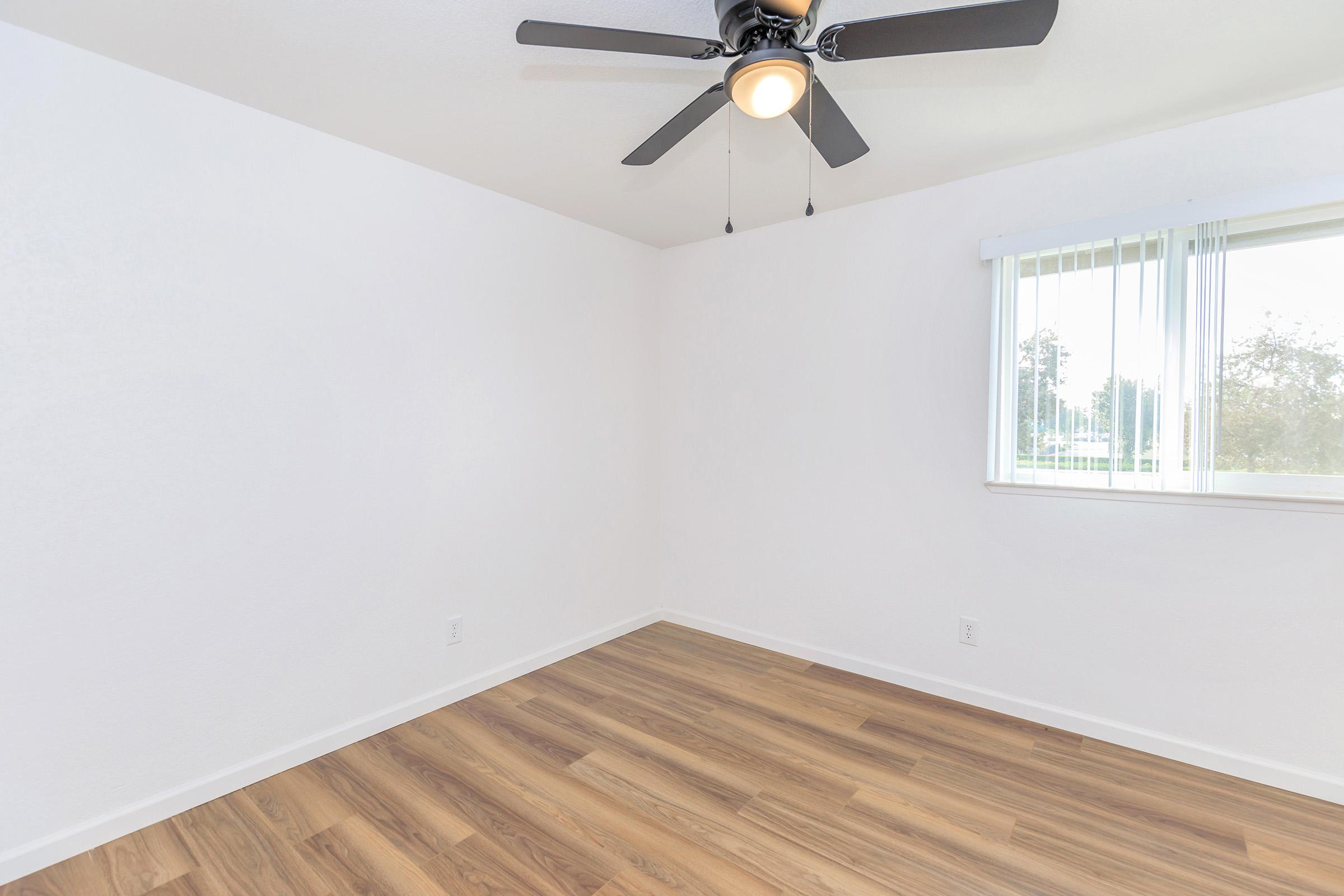 Empty room with light-colored walls and wooden flooring. A ceiling fan with three blades hangs from the ceiling. There is a window with vertical blinds allowing natural light to enter the space. The room appears bright and spacious, suitable for various uses or furnishings.
