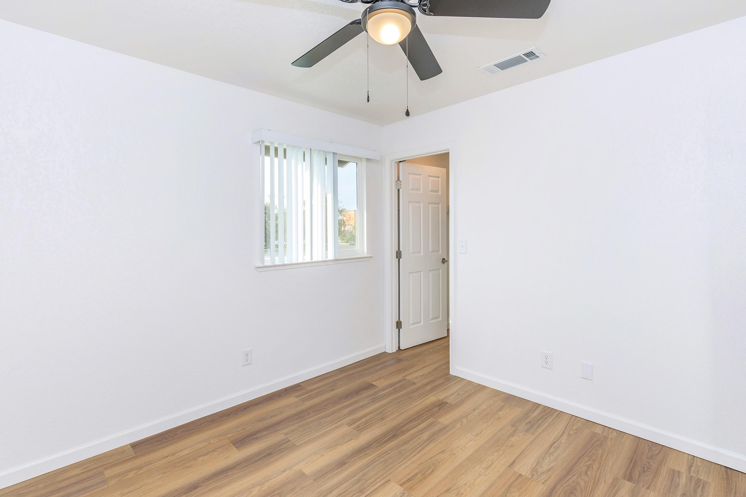 A bright, empty room featuring light-colored walls and wooden flooring. A ceiling fan is installed, and there is a window with vertical blinds allowing natural light. A closed door is visible, leading to another area of the space. The overall ambiance is clean and modern.