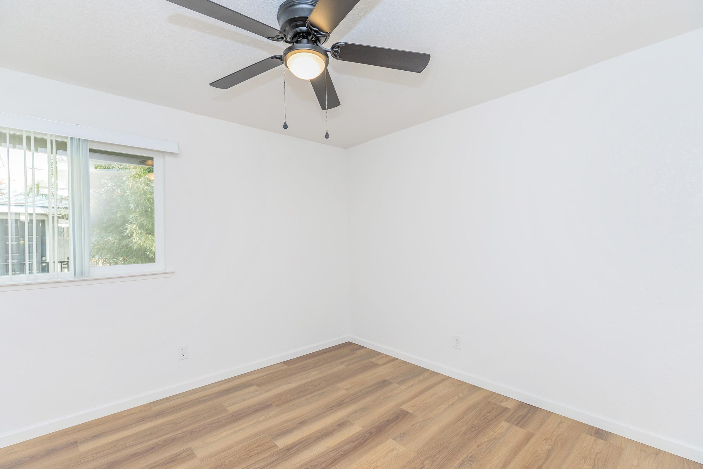 Empty room featuring light-colored walls and a wooden floor. A ceiling fan with a light fixture is mounted in the center. A window with vertical blinds allows natural light to enter. The space appears clean and well-maintained, suitable for various uses or decoration.