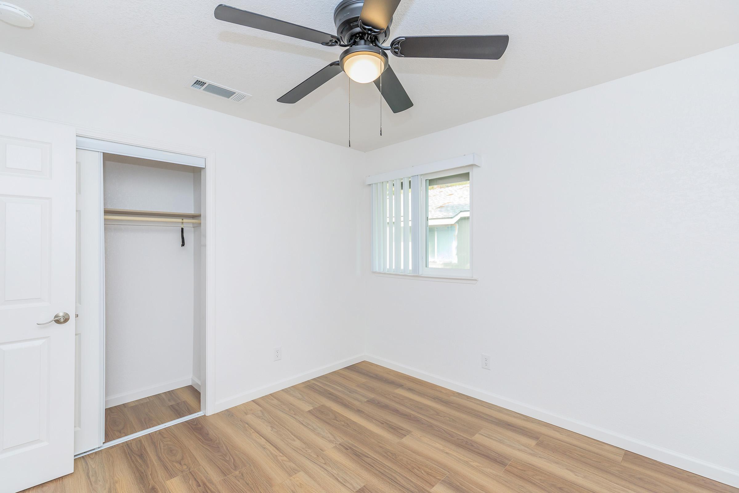 Empty room with light-colored walls and a ceiling fan. There is a window with blinds, a built-in closet with a shelf, and wooden flooring. The overall appearance is modern and minimalist.