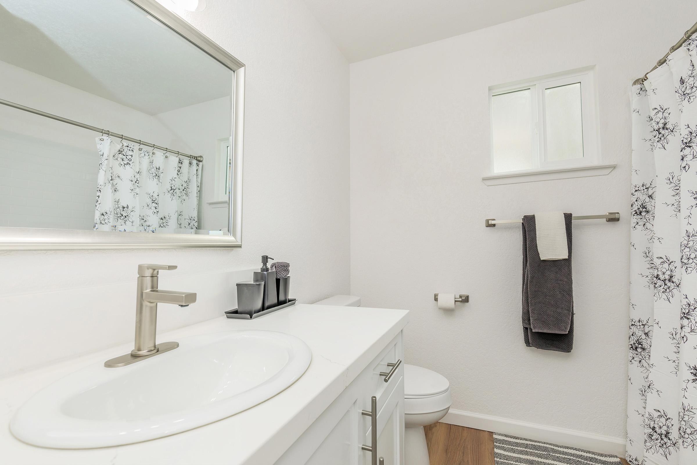 A modern bathroom featuring a white vanity with a sink, a large mirror, a white toilet, and a shower with a floral curtain. The walls are painted in a light color, and there are small windows providing natural light. A towel rack holds a gray towel, and there is a decorative soap dispenser on the counter.