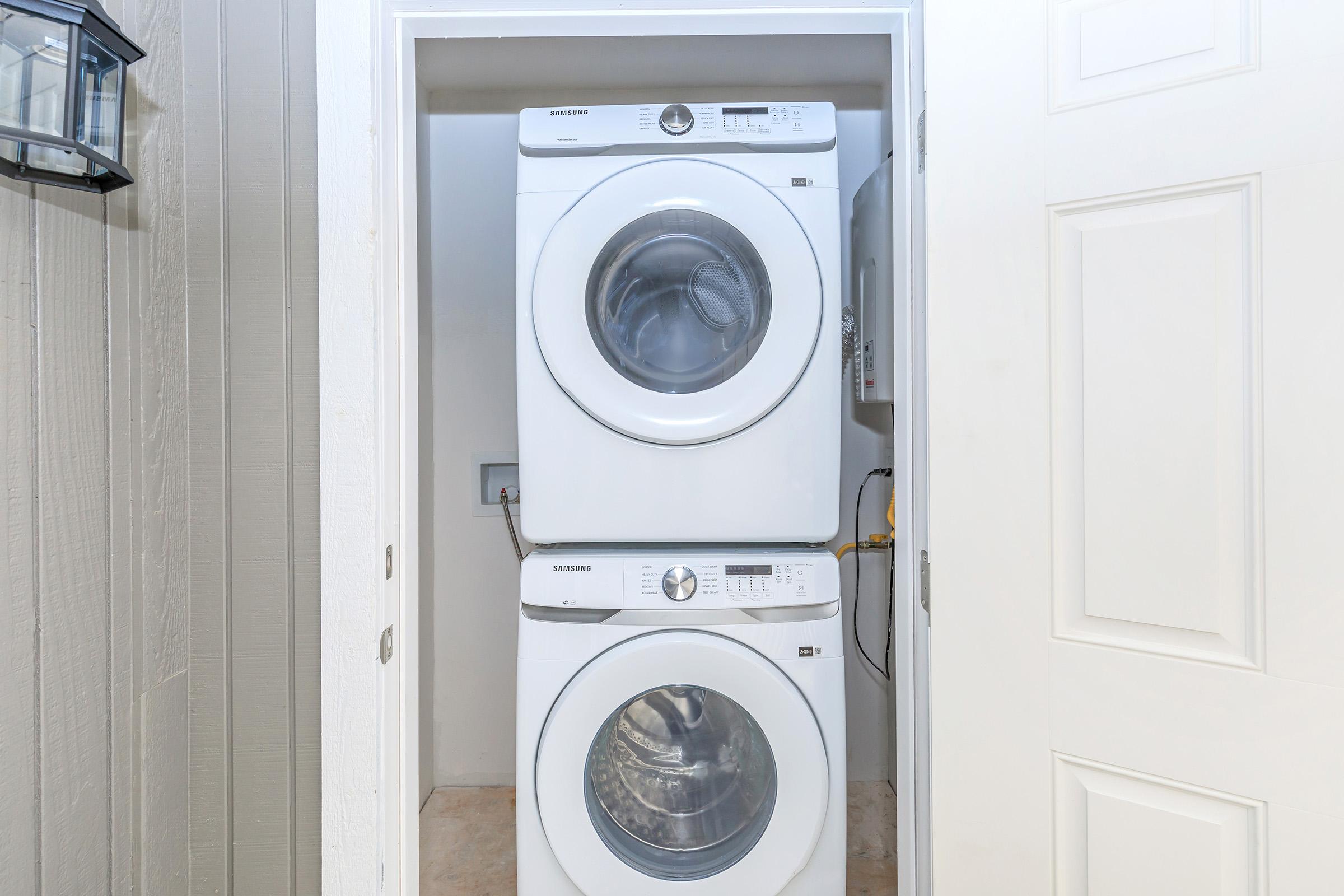 A stacked washer and dryer in a compact laundry closet, featuring a white finish and clear glass doors. The space has light-colored walls and a door on the right, suggesting a tidy and organized laundry area.